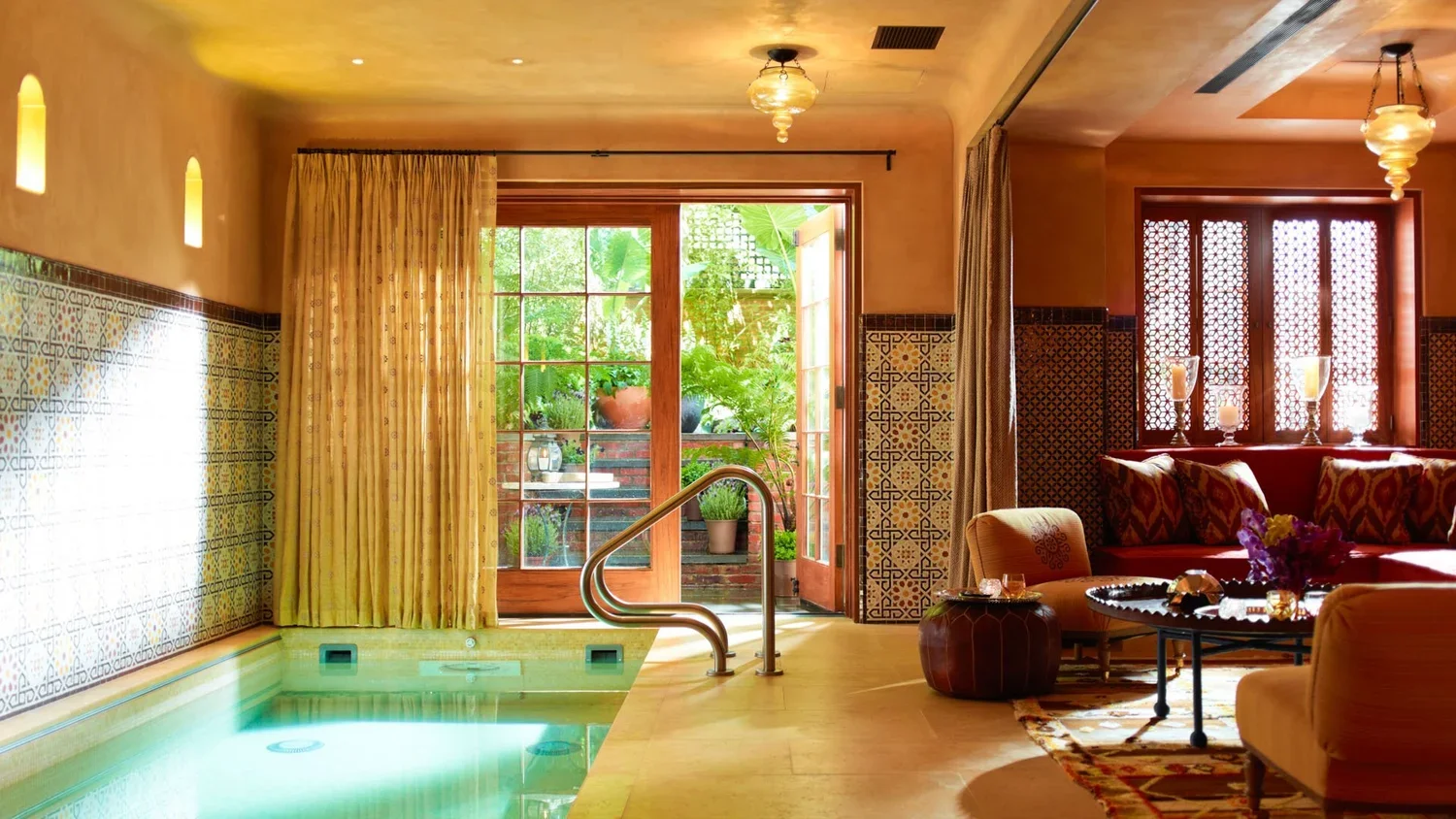 Luxurious indoor room with a hot tub, floral and geometric patterned tiles, large windows with curtains, and cozy seating area with pillows and candles, opening to a garden with potted plants.