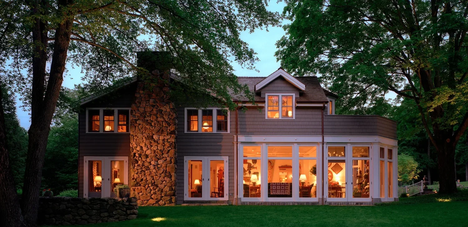 A two-story house with large windows, warm interior lighting, a stone chimney, surrounded by green trees and a well-maintained lawn.