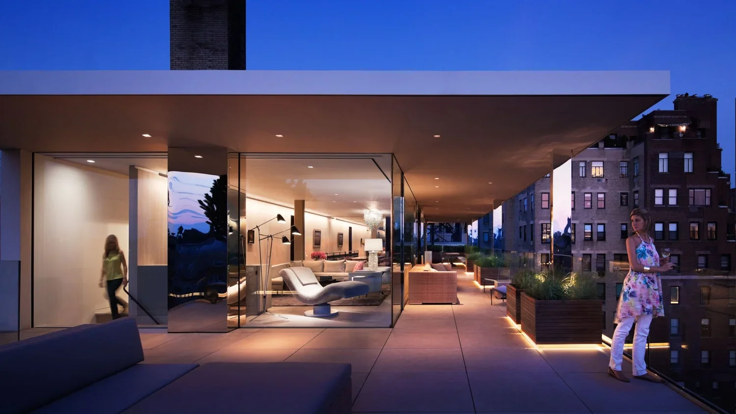 Nighttime view of a modern apartment balcony with outdoor seating, potted plants, and two women, one standing and one sitting, overlooking city buildings.