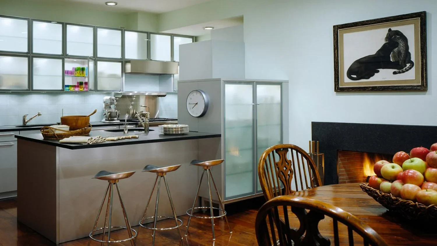 Modern kitchen with an island, bar stools, a fireplace, and a dining table with a basket of apples. There's a framed animal artwork on the wall.