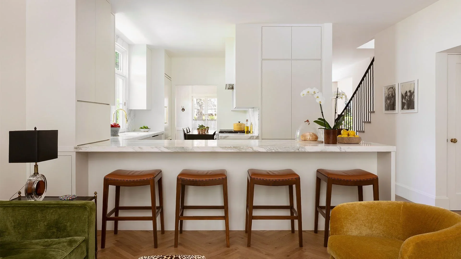 Modern kitchen with white cabinets, marble countertops, and a breakfast bar with four brown stools. There are vases with flowers and fruit on the counter, and a green velvet chair with a black lamp beside it in the foreground.
