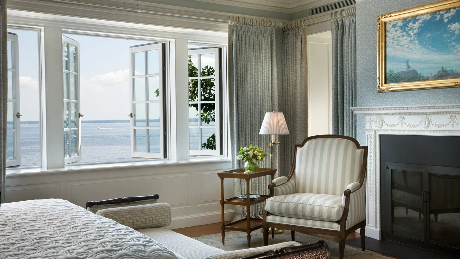 A bedroom with large open windows overlooking a body of water and a boat. The room has a striped upholstered armchair, a side table with a vase of flowers, a lamp, and a framed landscape painting above the fireplace.