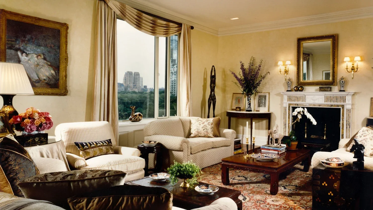 Elegant living room with cream-colored couches, dark wood coffee table, fireplace with mirror, framed art, end tables with lamps and decorative items, window with curtains, and various decorative objects and flowers.