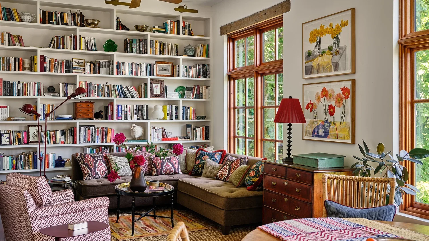 Living room with a white bookshelf filled with books, decorative items, and framed pictures. Large windows with wooden trim let in natural light. A beige sectional sofa with colorful pillows is centered with a black round coffee table holding a vase 