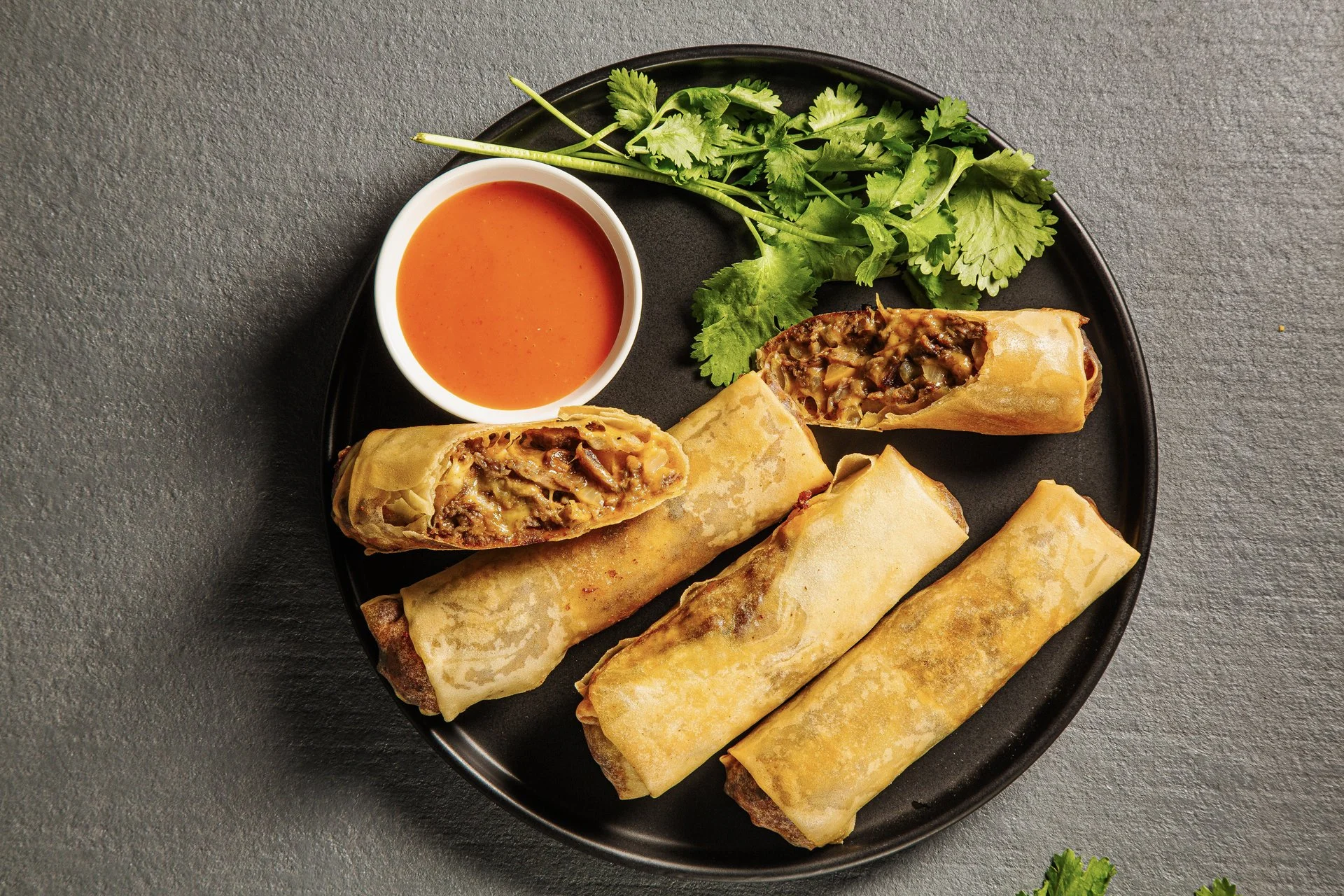 Plate with four egg rolls, a small bowl of orange dipping sauce, and fresh cilantro on a dark gray surface.