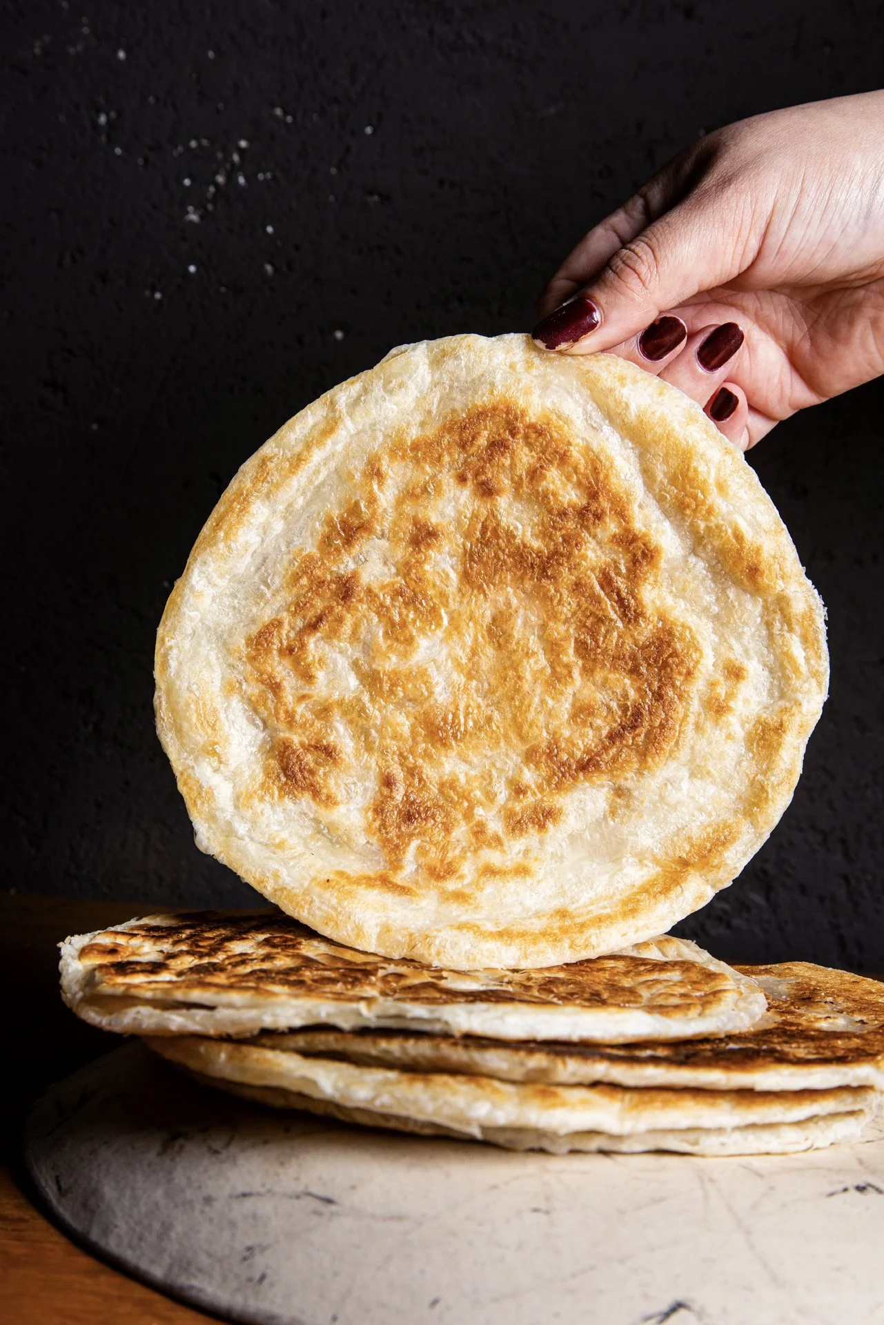 A hand holding a cooked flatbread or pancake over a stack of similar cooked bread on a marble surface.