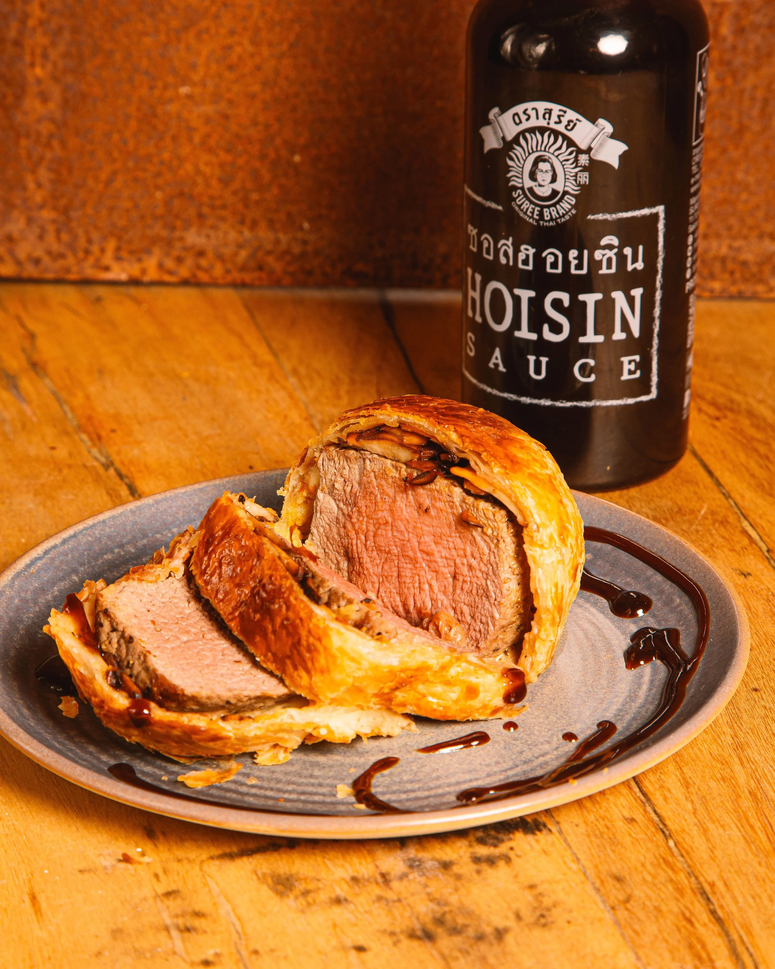 A plate with a beef Wellington sliced open, showcasing the cooked beef and pastry crust. The Wellington is drizzled with hoisin sauce, which is also on a bottle in the background labeled "Hoisin Sauce".