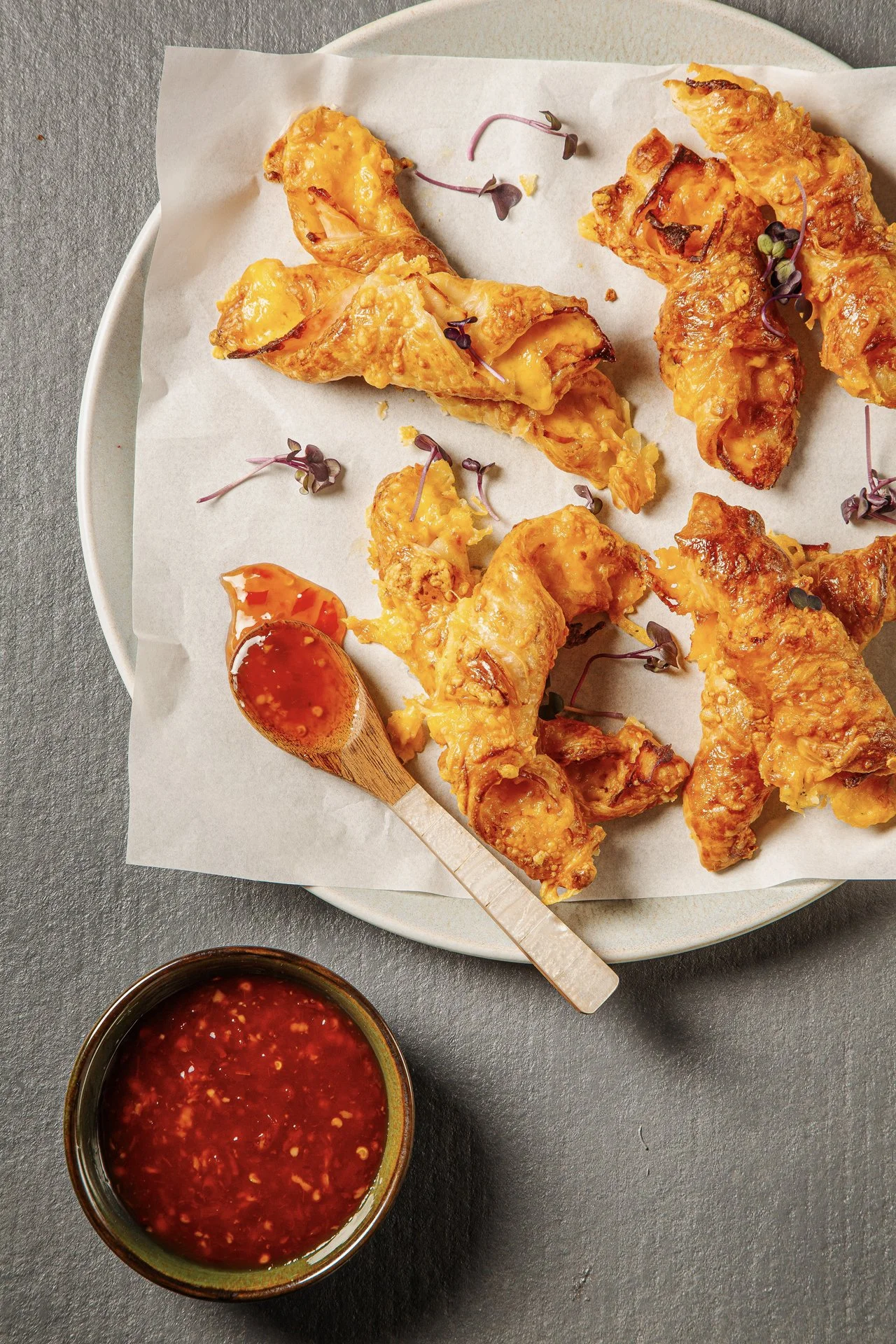 Fried chicken wings with a side of red chili sauce, garnished with microgreens, served on a white plate with parchment paper, on a gray surface.