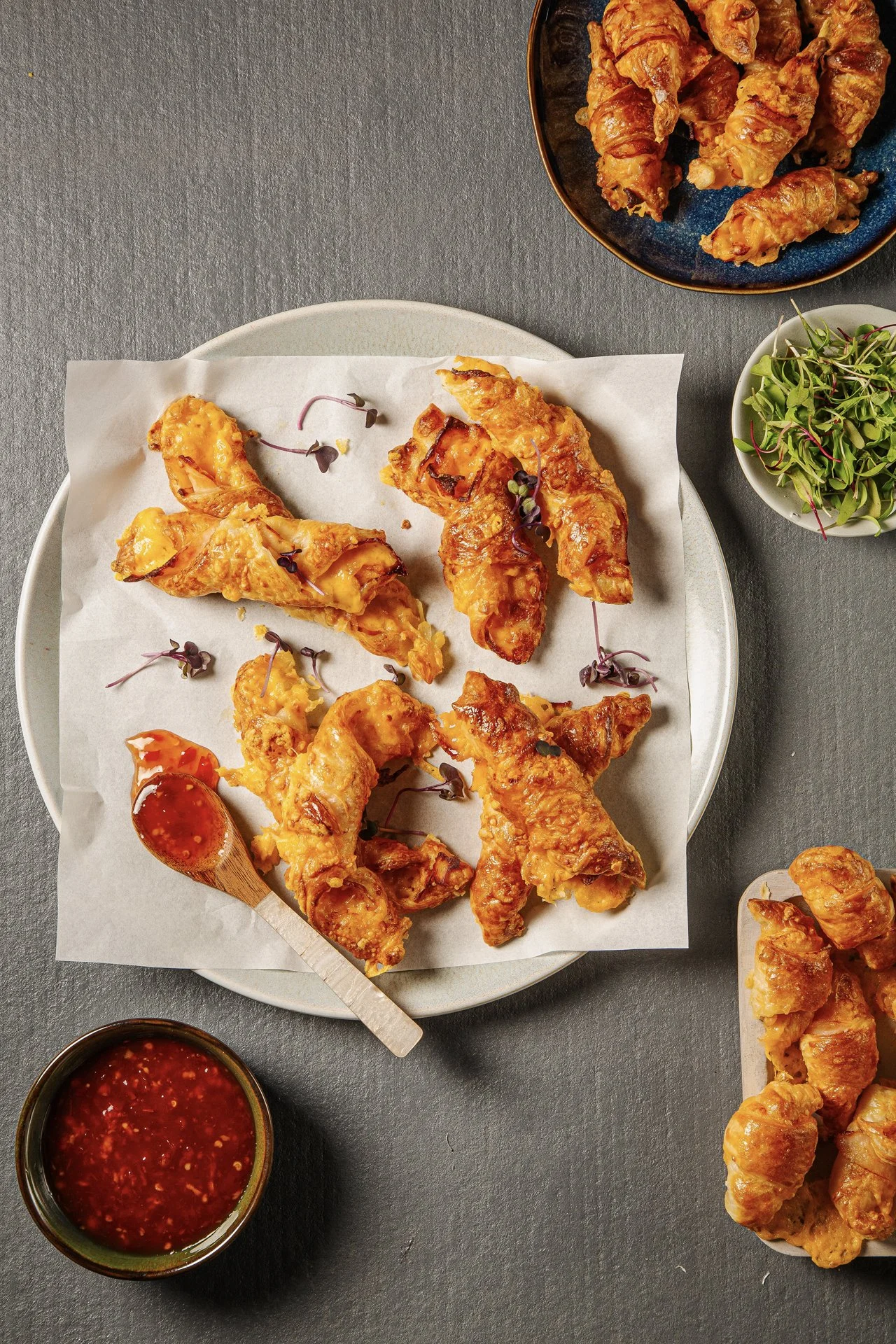 A top-down view of a plate of fried chicken wings with microgreens, a small bowl of dipping sauce, and additional fried chicken pieces, with some side salad leaves and more fried chicken in bowls.