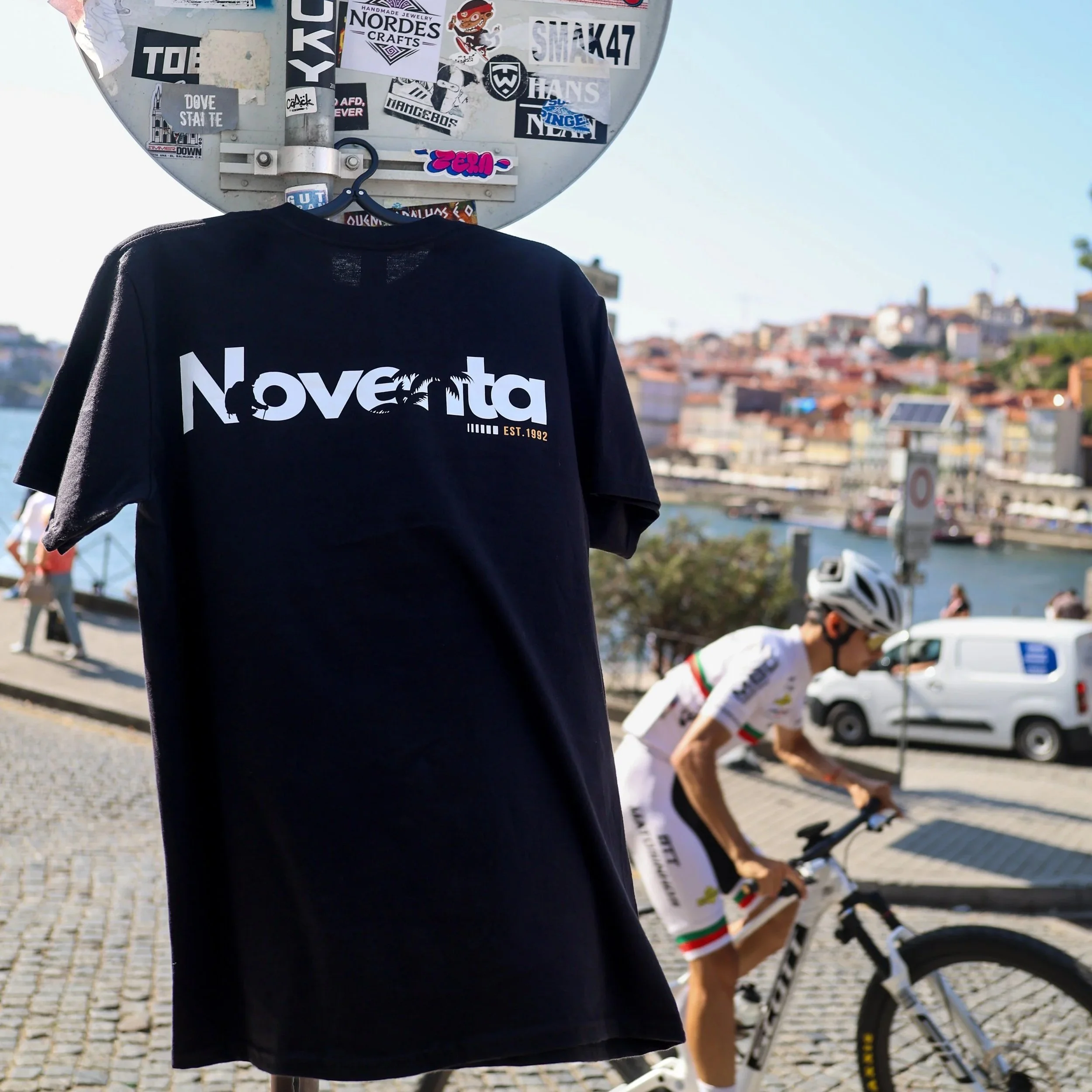 A black T-shirt hanging on a board with a sticker in the background and a cyclist riding a white bike on a cobblestone street near a river and colorful buildings.
