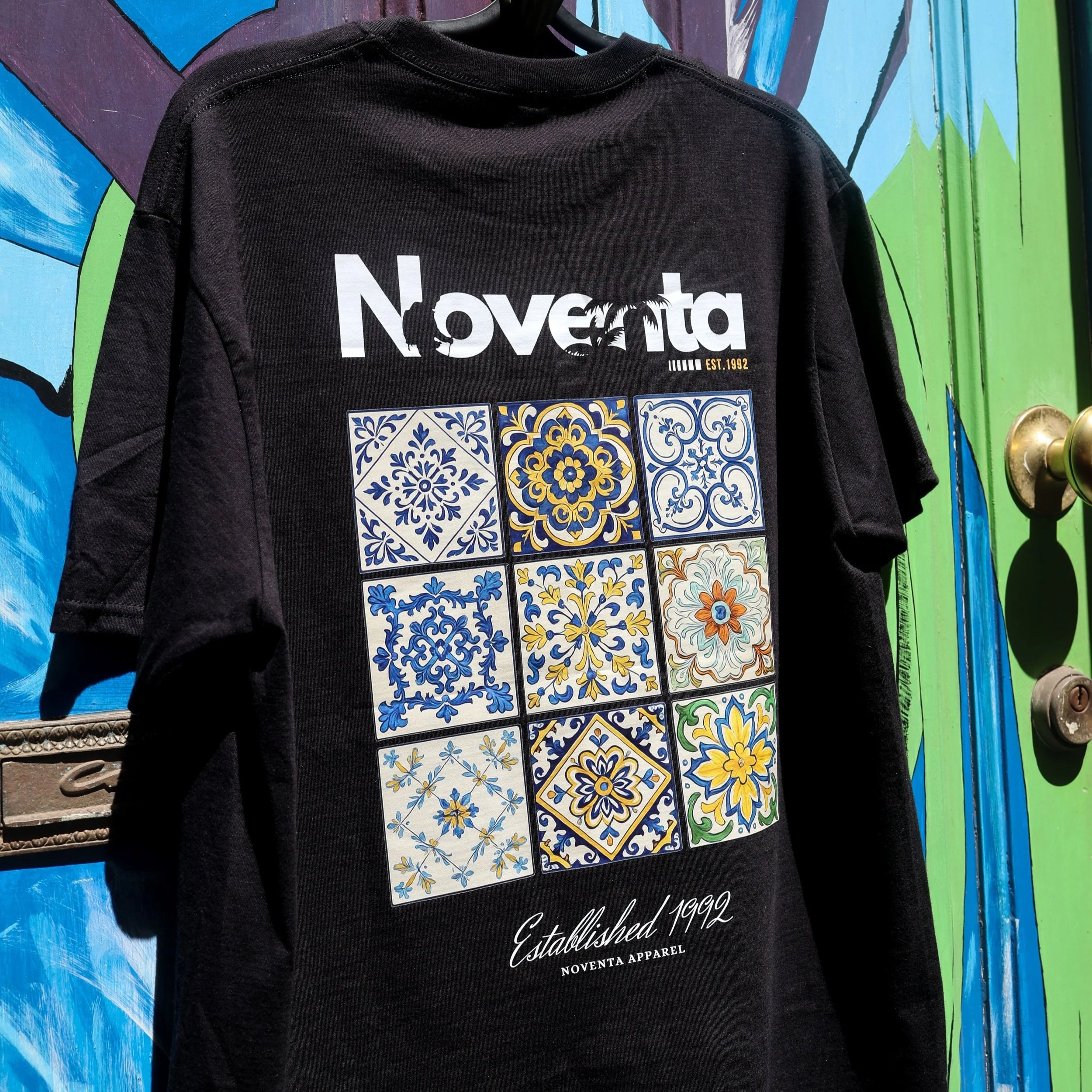 Black t-shirt with white 'Noventa' text, nine decorative tile patterns in a 3x3 grid, and script 'Established 1992' at the bottom, hanging in front of a colorful wall.