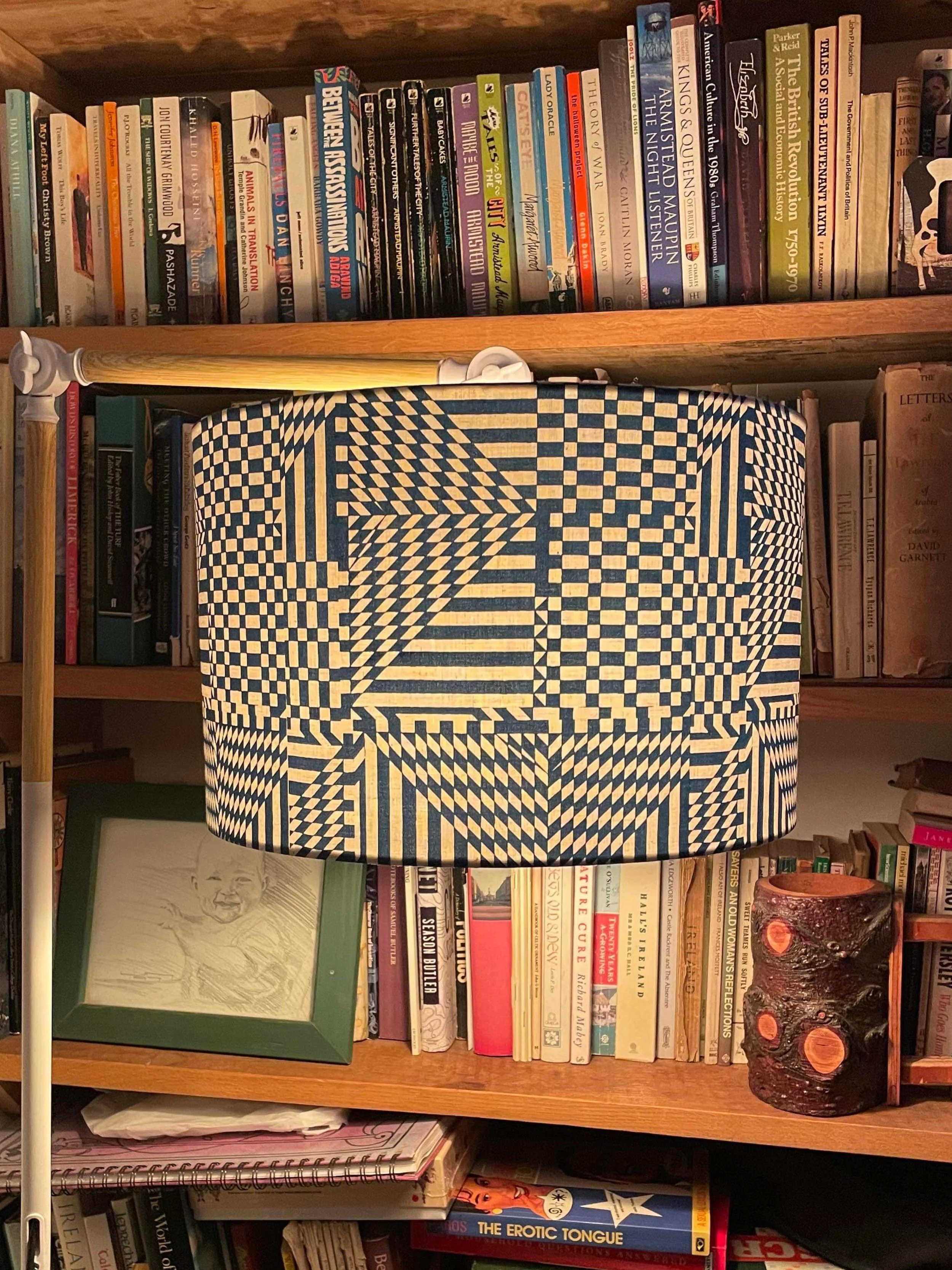 A geometric patterned lampshade with black and white angular designs hanging in front of a bookshelf filled with books, framed art, and decorative objects.