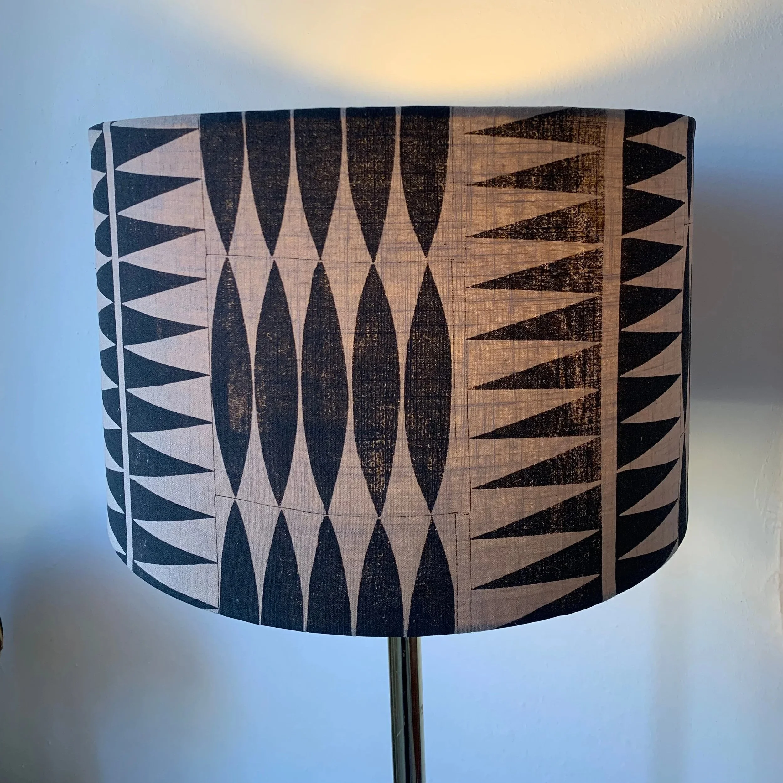 A lampshade with a black geometric pattern of elongated oval and triangular shapes on a light background, mounted on a brass-colored lamp base.