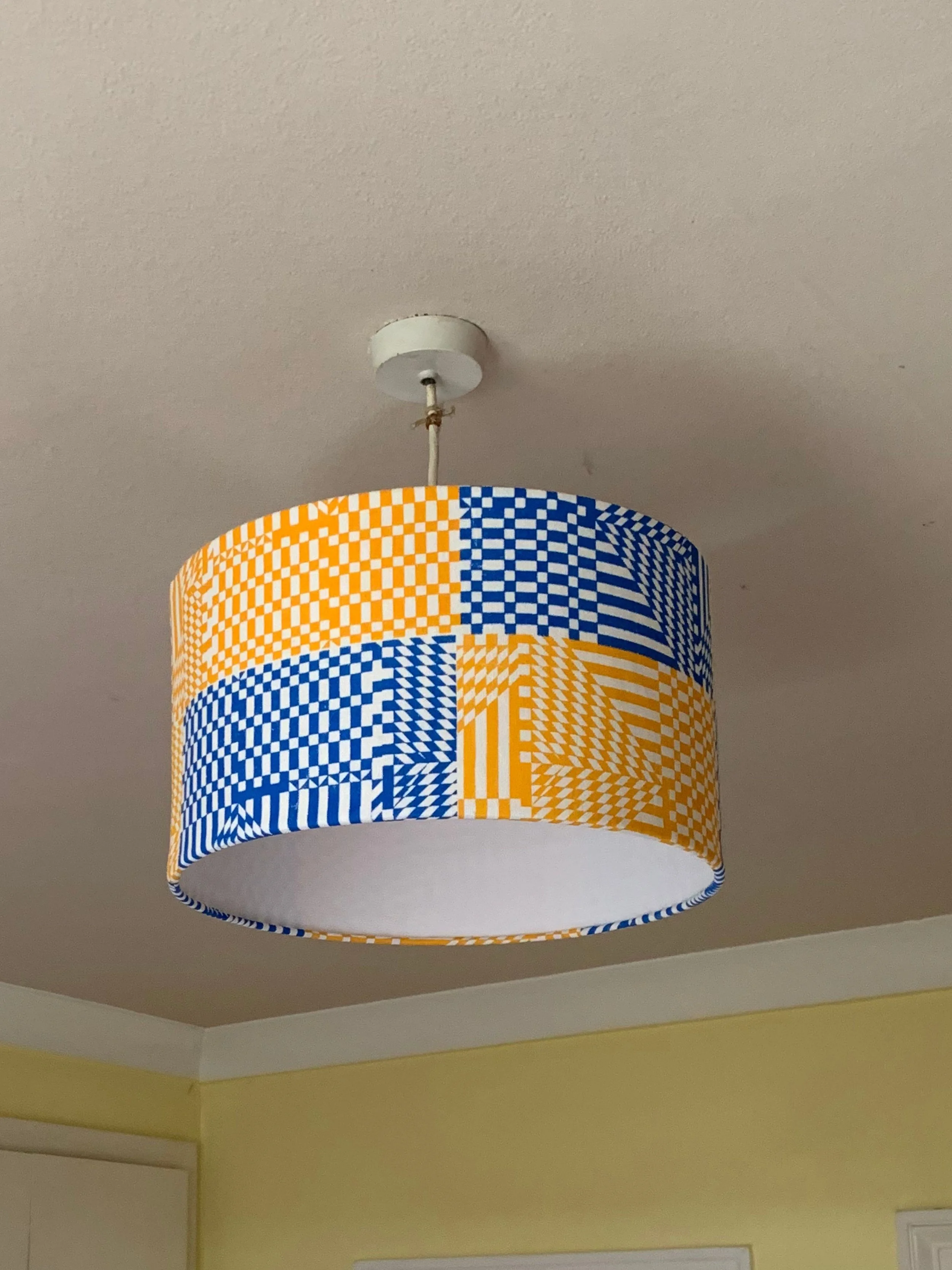 A ceiling light fixture with a colorful patterned lampshade in blue, orange, and white geometric designs, hanging from a white ceiling in a room with yellow walls.