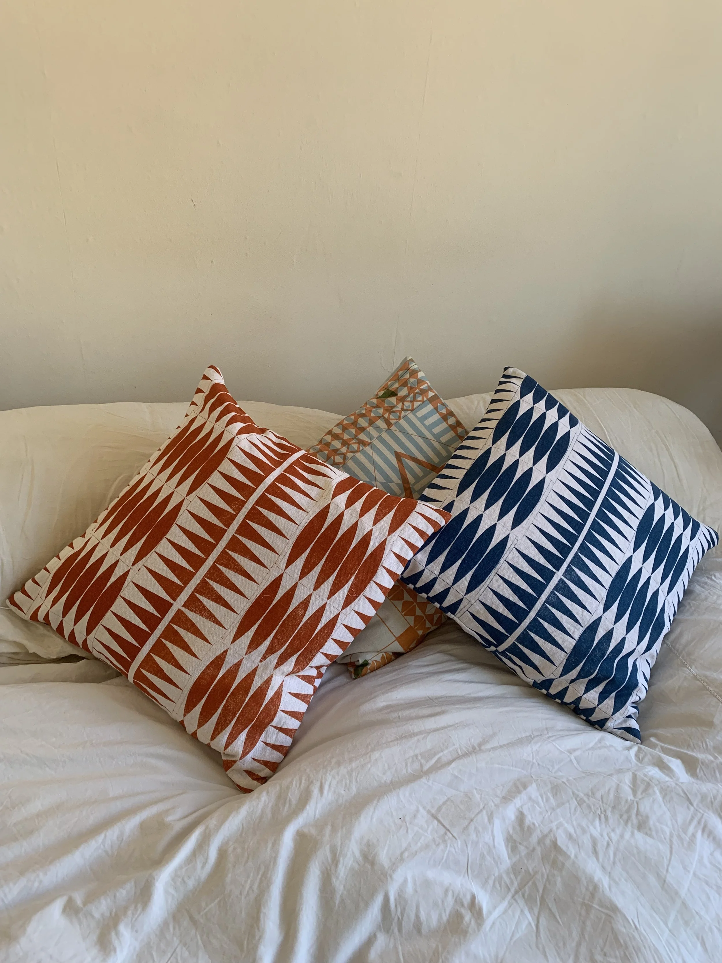 Three decorative throw pillows with geometric patterns in red, orange, and blue placed on a white bed against a plain wall.