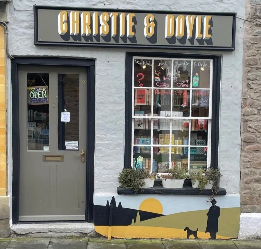 Christie and Doyle shop front. Graphic sign of shop's name. Painting of hills, a detective, and a small dog.