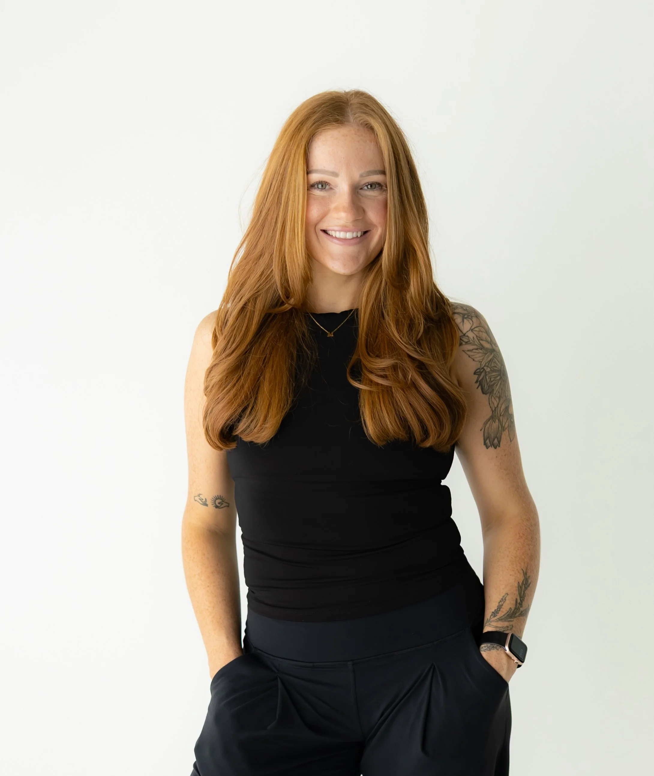Young woman with long red hair, wearing a black t-shirt and beige pants, sitting against a plain white background, smiling with her arms crossed.