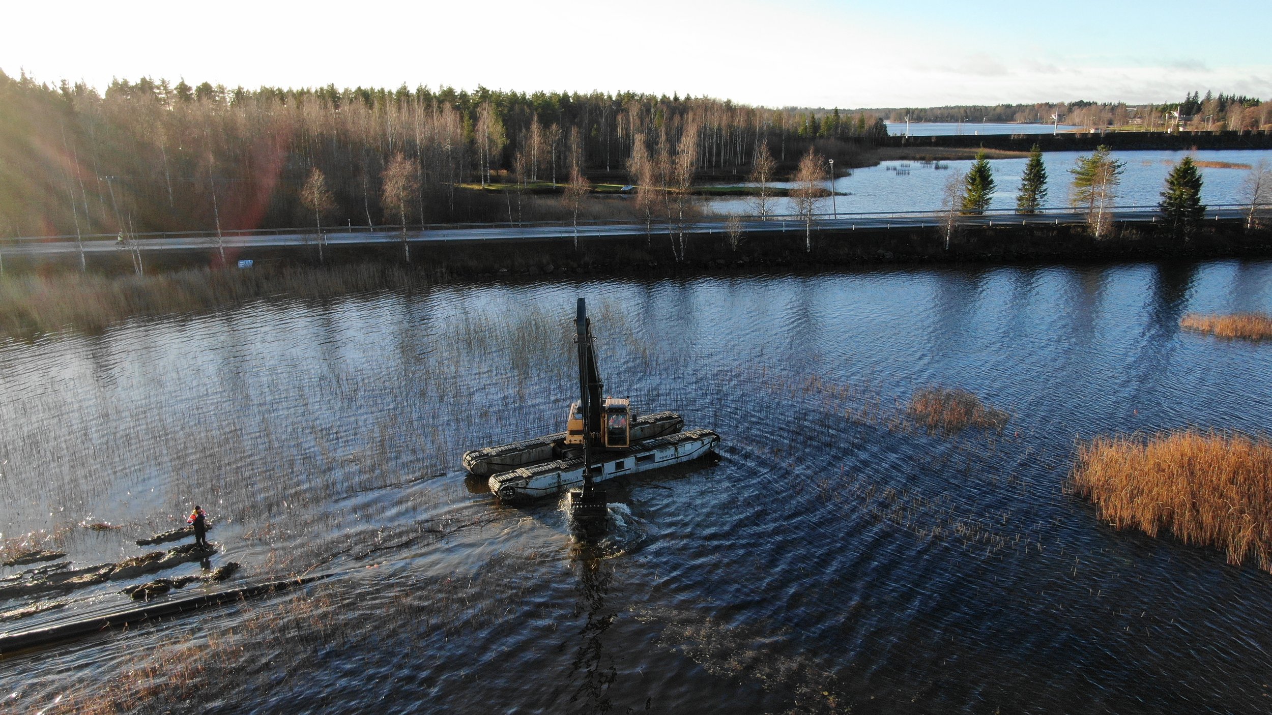 Ilmakuva suuresta ponttoonikoneesta, joka on asennettu veteen, ja miehestä menossa kohti laitetta. Taustalla on joki, tie ja metsää puita.