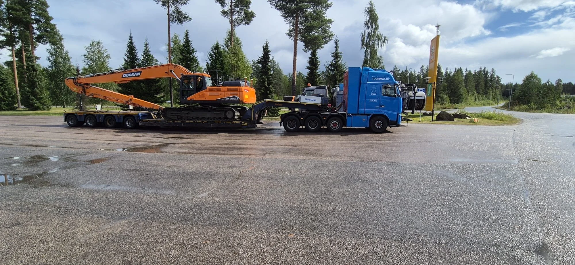 Sinisyksellä on iso sininen kuorma-auto, johon on lastattu pieni oranssi kaivuuromu. Taustalla on metsä ja taivas, ja tiellä on pieni lätäkkö.