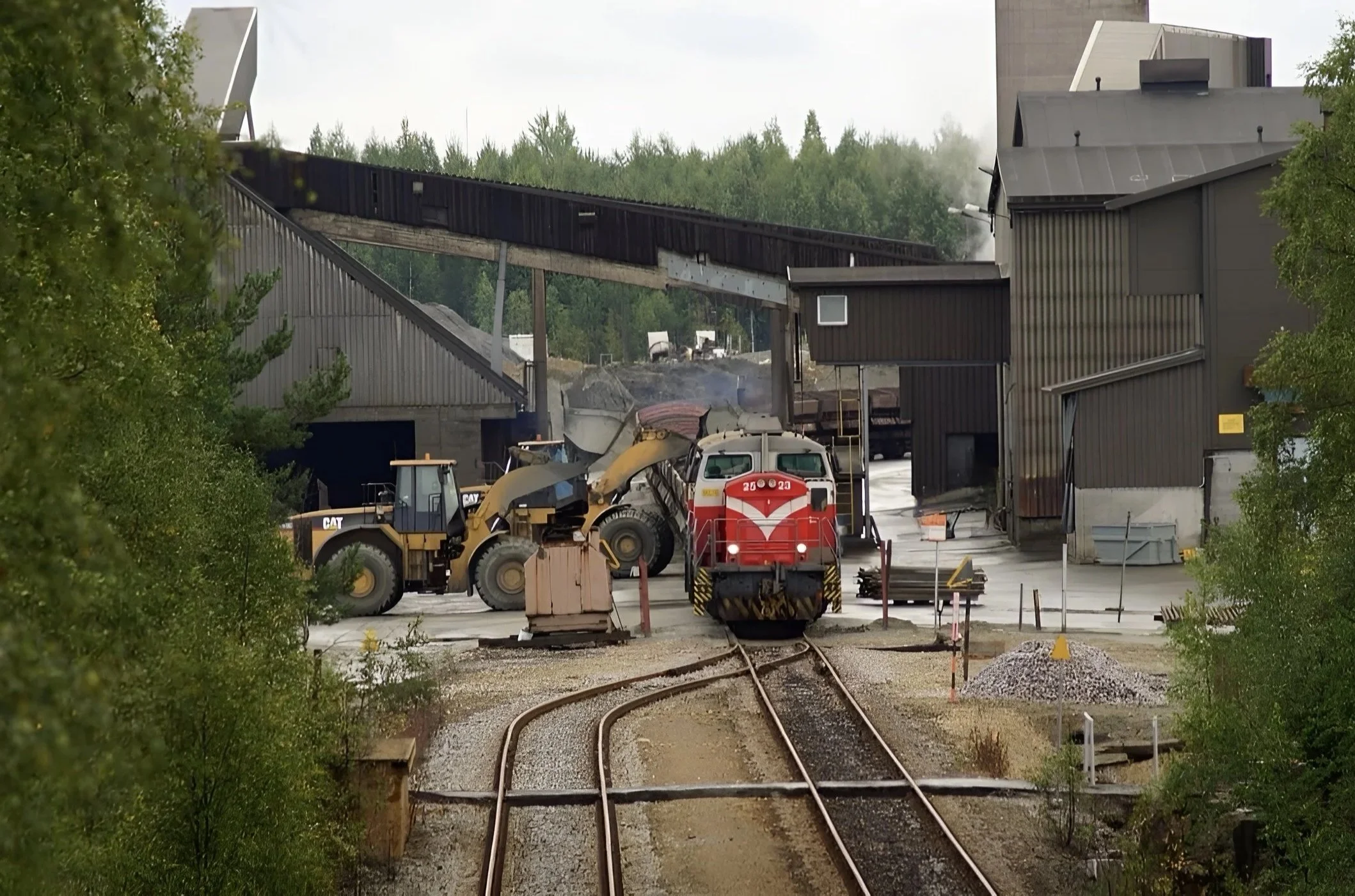 Kuvassa rakennustyömaa veturilla ja kaivukoneella, jotka sijaitsevat rautatiealueen keskellä, ja ympärillä on puita ja teollisuusrakennuksia.