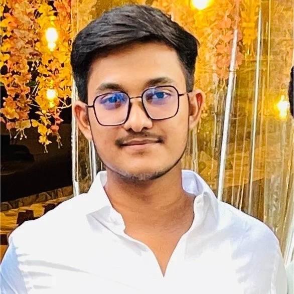 A young man with glasses and short dark hair, wearing a white shirt, standing in front of a decorative background with yellow flowers and warm lighting.