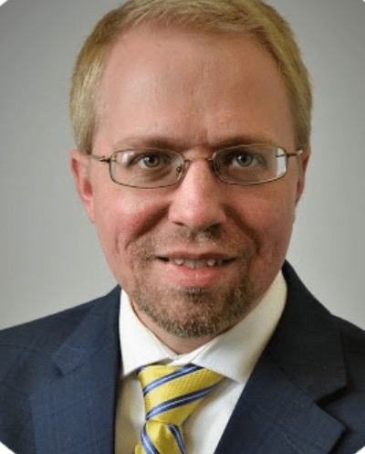 Close-up of a man with glasses, light-colored hair, wearing a dark suit, white shirt, and yellow striped tie, smiling against a neutral background.
