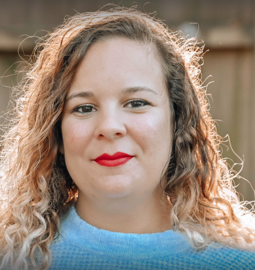 A woman with curly, shoulder-length hair and red lipstick, wearing a blue sweater, smiling outdoors with sunlight behind her.