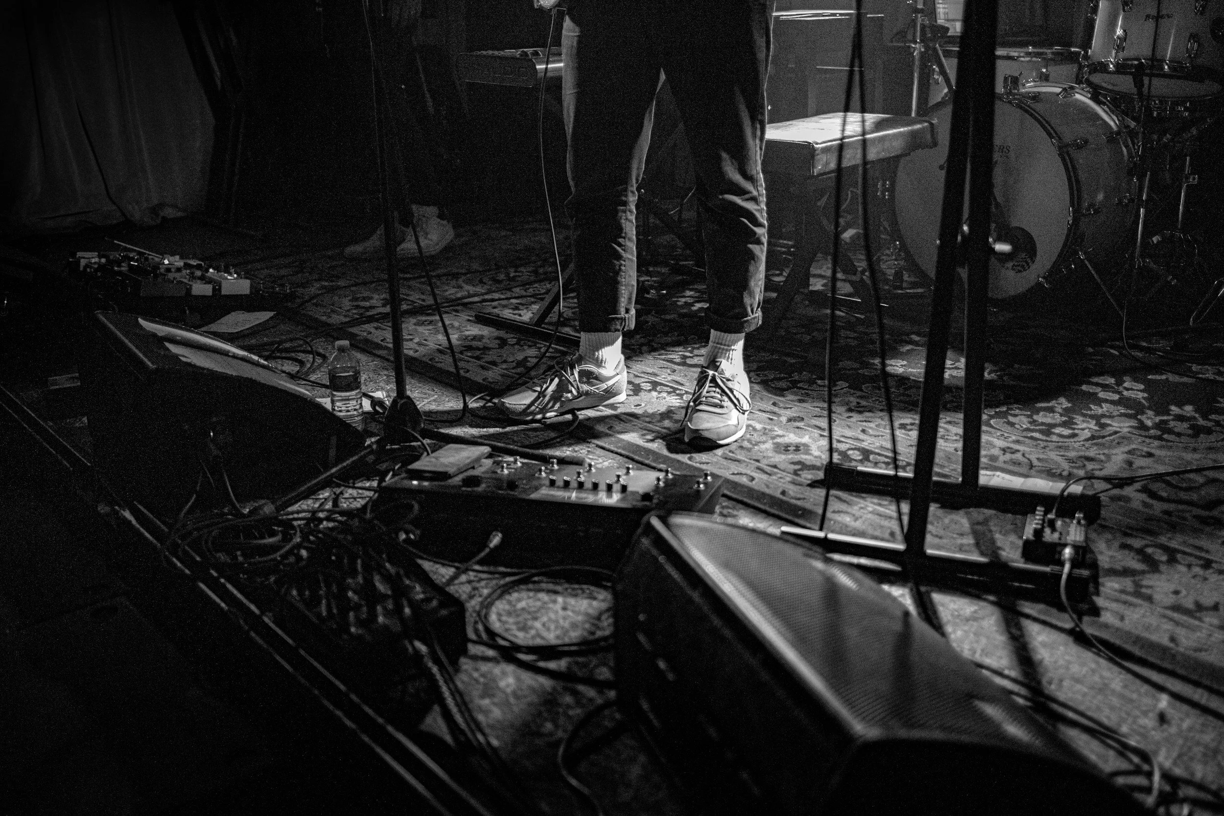 Black and white photo of a musician on stage, showing only their lower body, standing in front of musical equipment, drums, pedals, and a water bottle, with the stage decorated by an ornate rug.