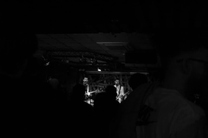 A live music performance in a dark venue with two guitarists on stage, illuminated by sparse lighting, facing an audience.