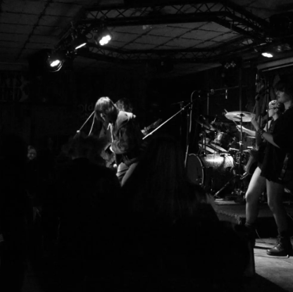 Musicians performing on a stage with drums and microphones in a dark venue.