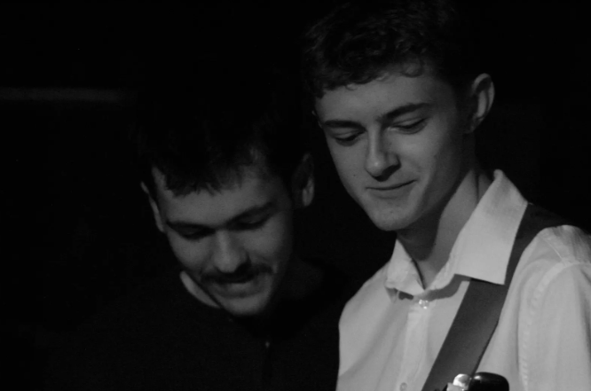 Two young men are closely together, looking down and smiling in a dark setting.