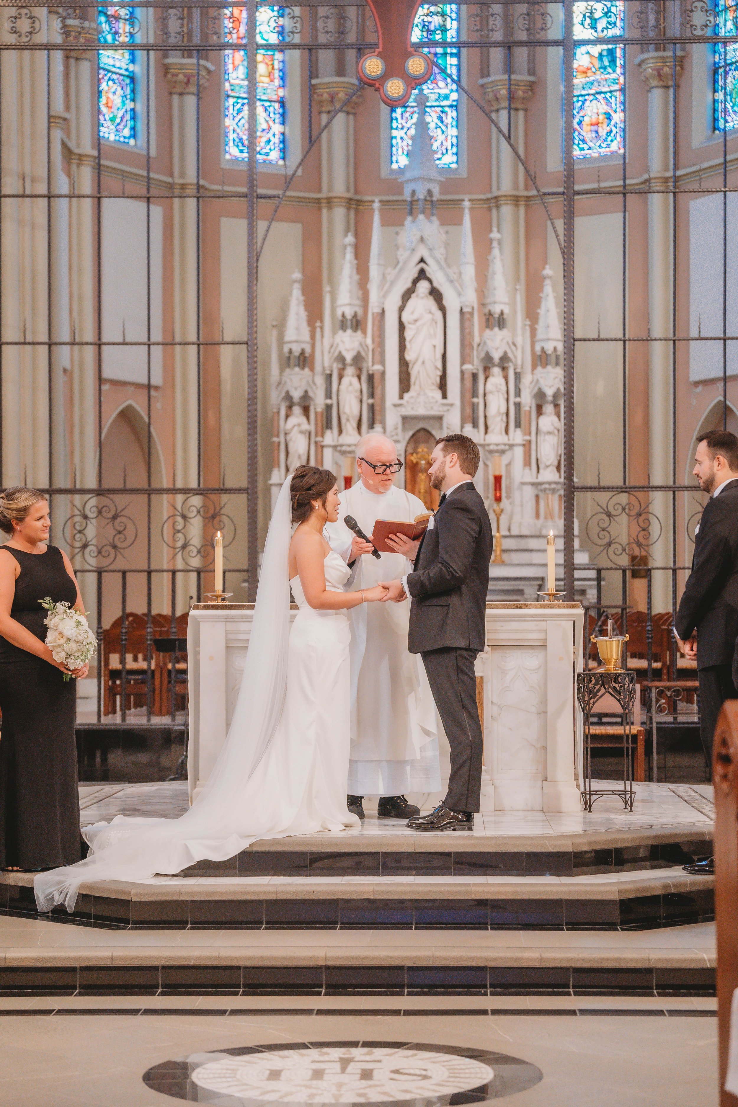 A wedding ceremony taking place in a church with stained glass windows. The bride and groom hold hands and face each other before a priest. The bride is in a white gown with a long veil, and the groom is in a black suit. A bridesmaid holds a bouquet, and a groomsman stands to the side. The altar is decorated with candles and religious statues.