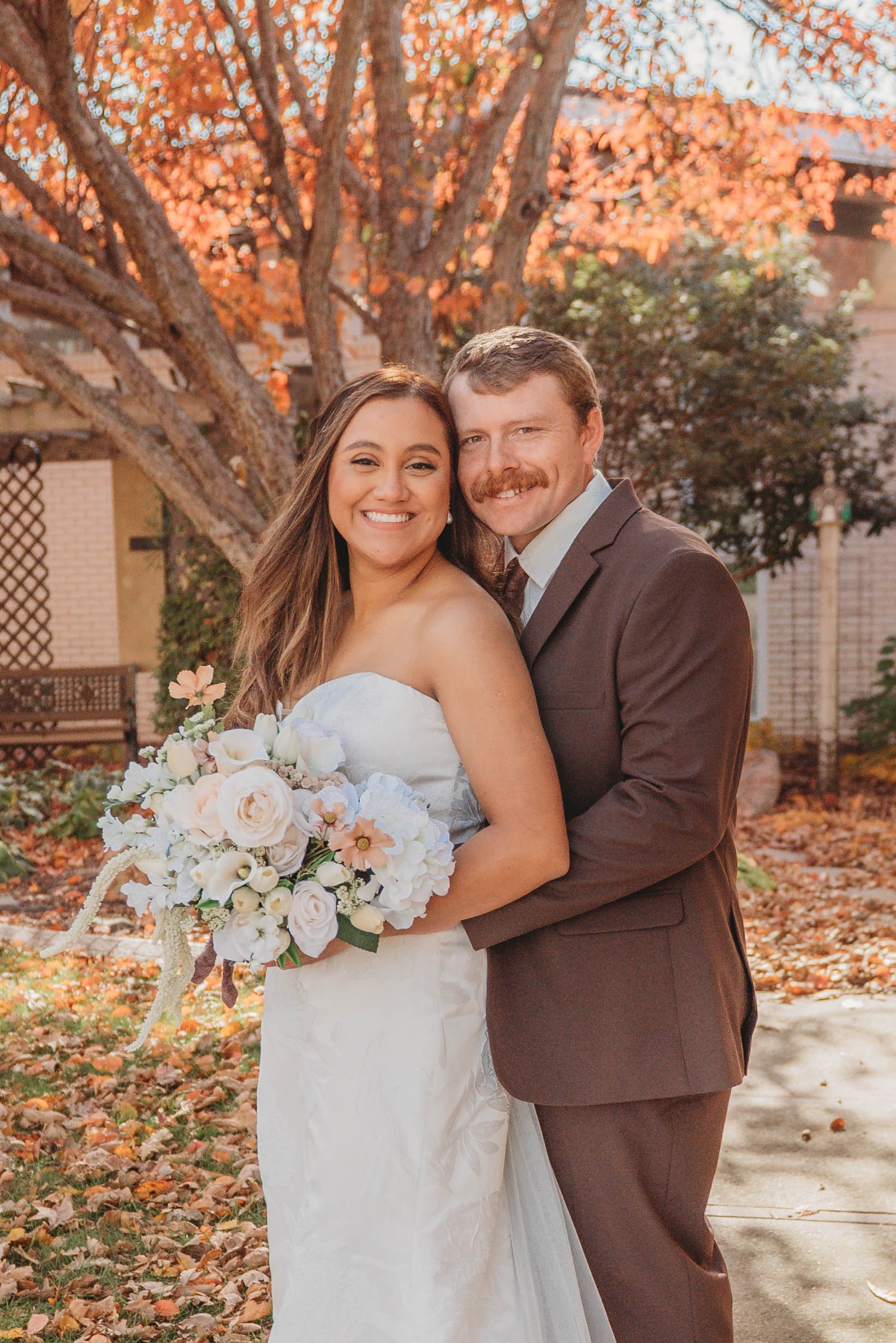 A smiling bride and groom in a garden during autumn, with orange leaves on trees in the background. The bride is holding a bouquet of white and pastel flowers, wearing a strapless white wedding dress. The groom is wearing a brown suit and has his arms around the bride.