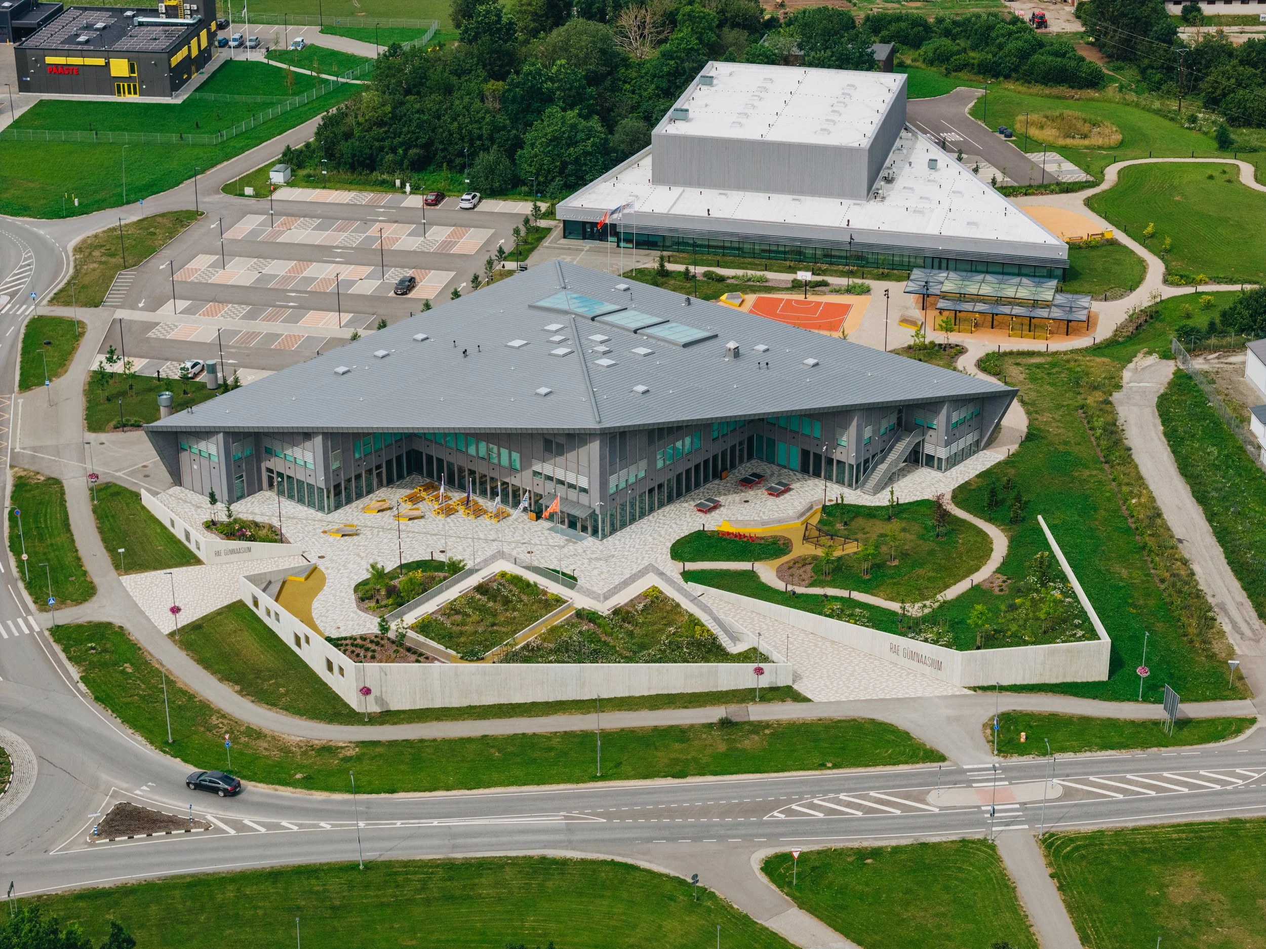 An aerial view of a modern school building with surrounding landscaped gardens, walkways, a parking lot, and sports facilities.