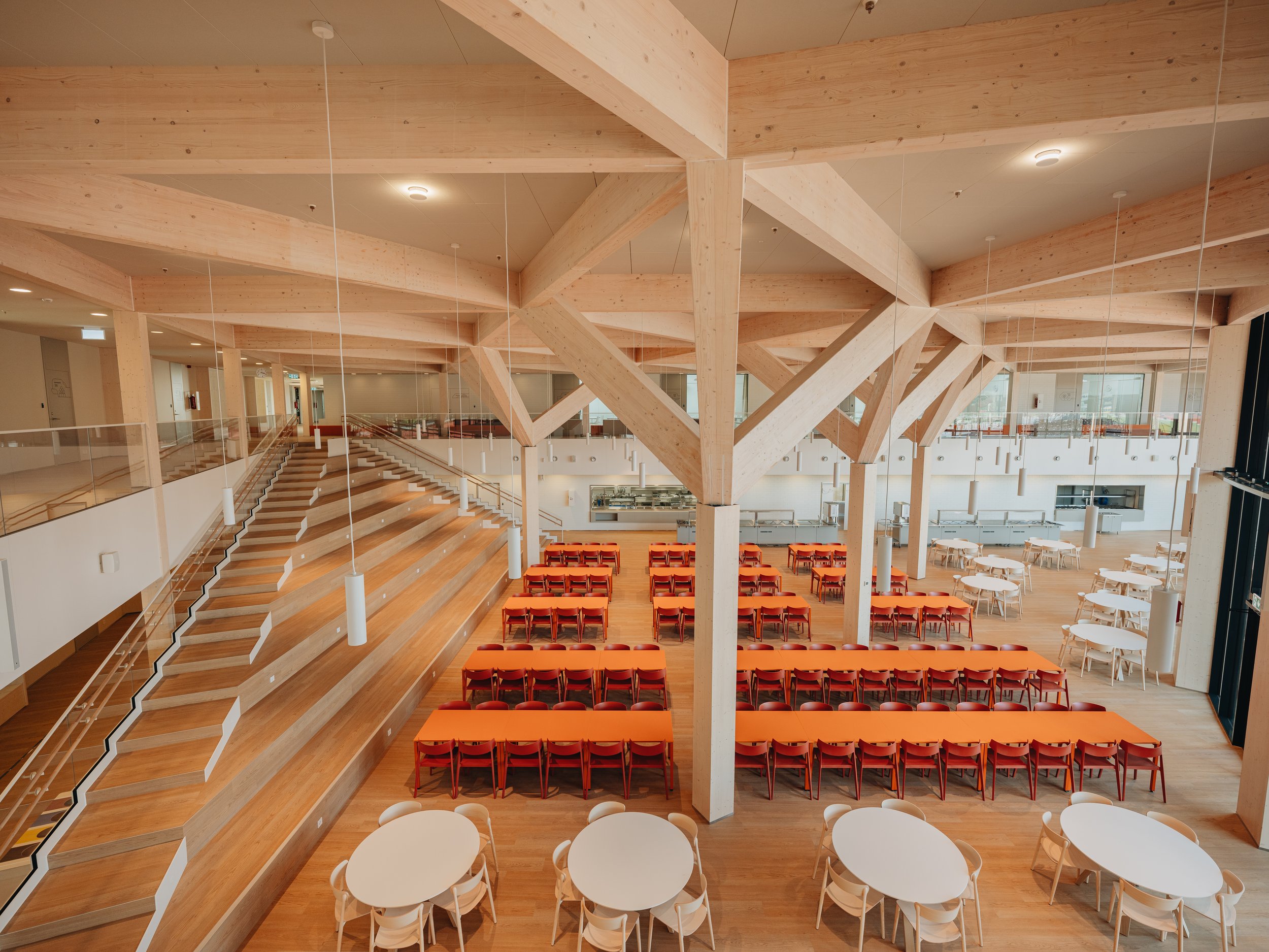 Interior view of a modern dining area with wooden beams, staircase, and tables with orange and white chairs.