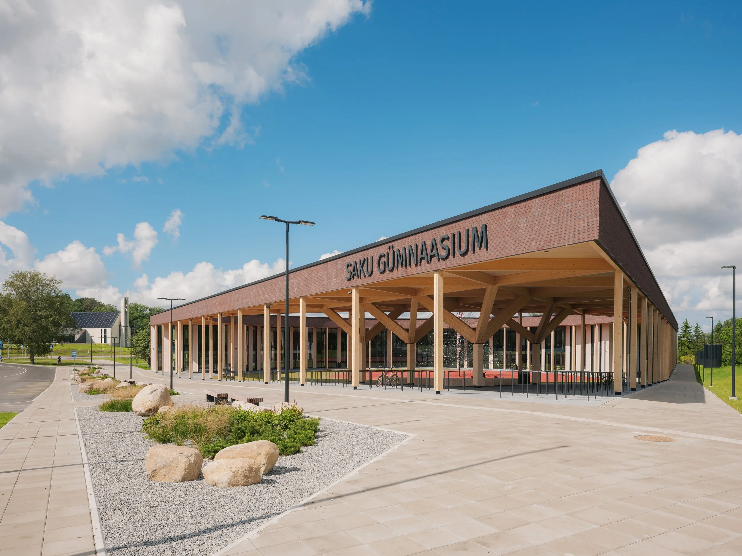 Exterior view of Saku Gymnasium, a modern building with a wooden support structure, brick upper exterior, and large glass windows, surrounded by a paved walkway, landscaping, and a partly cloudy sky.
