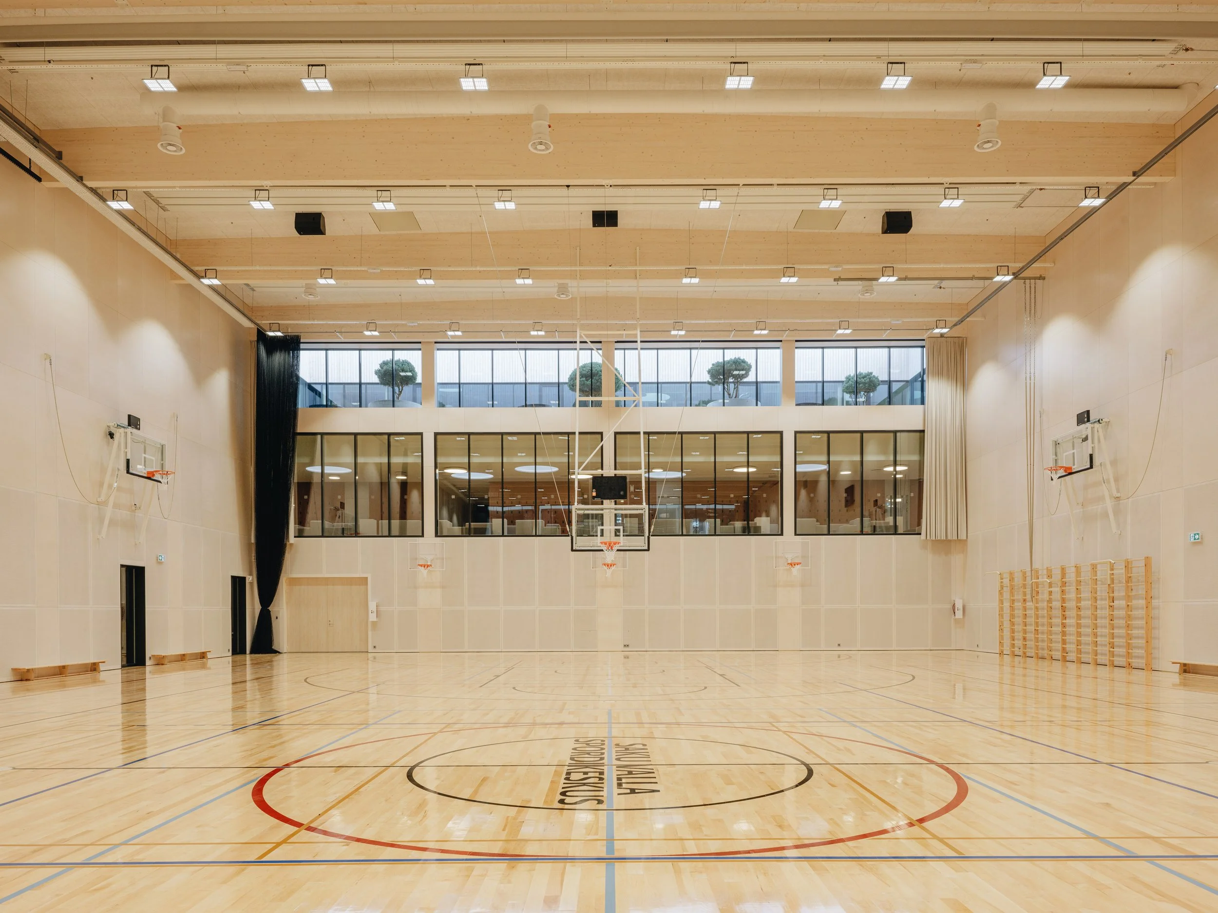 Empty indoor basketball court with wooden flooring, multiple basketball hoops on the walls, large windows at the back letting in natural light, and high ceilings with ceiling lights.