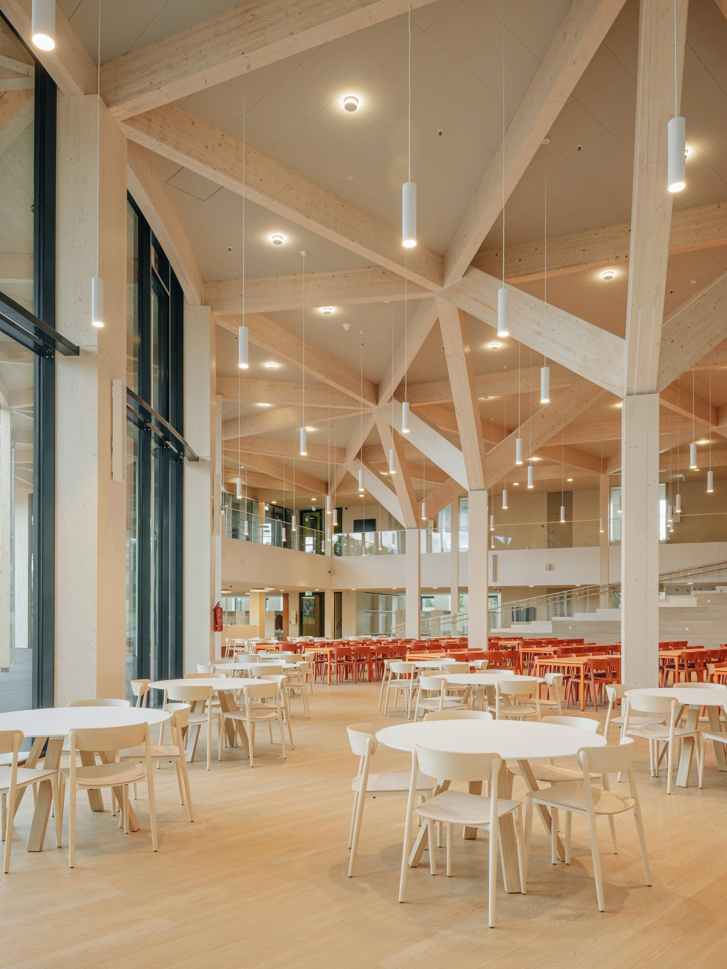 Spacious modern dining hall with wooden beams, pendant lighting, large windows, and white and orange tables and chairs.