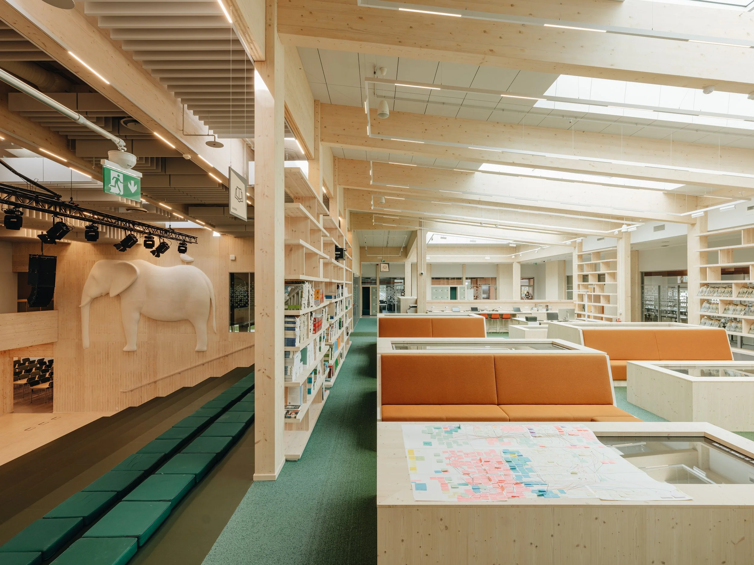Interior view of a modern, well-lit library with wooden accents, orange seating, bookshelves, and a large white elephant sculpture on a wall.