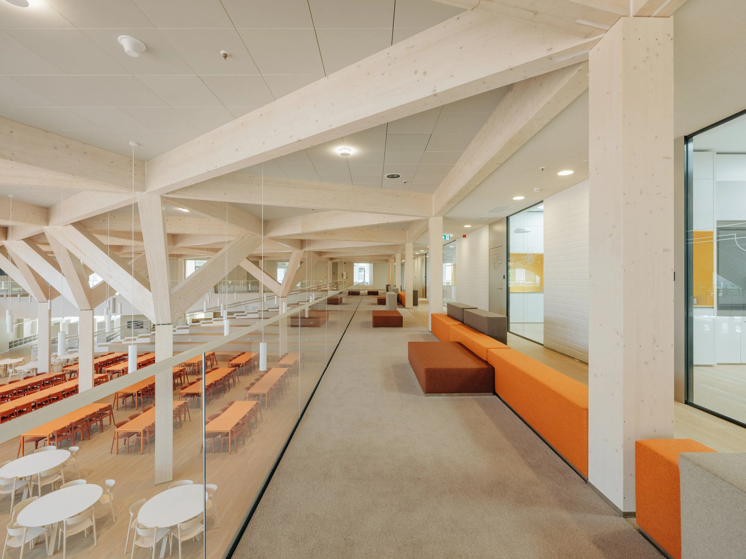 Modern indoor space with orange and gray seating along a glass wall, visible ceiling beams, and a dining area with rows of orange chairs and white tables underneath.