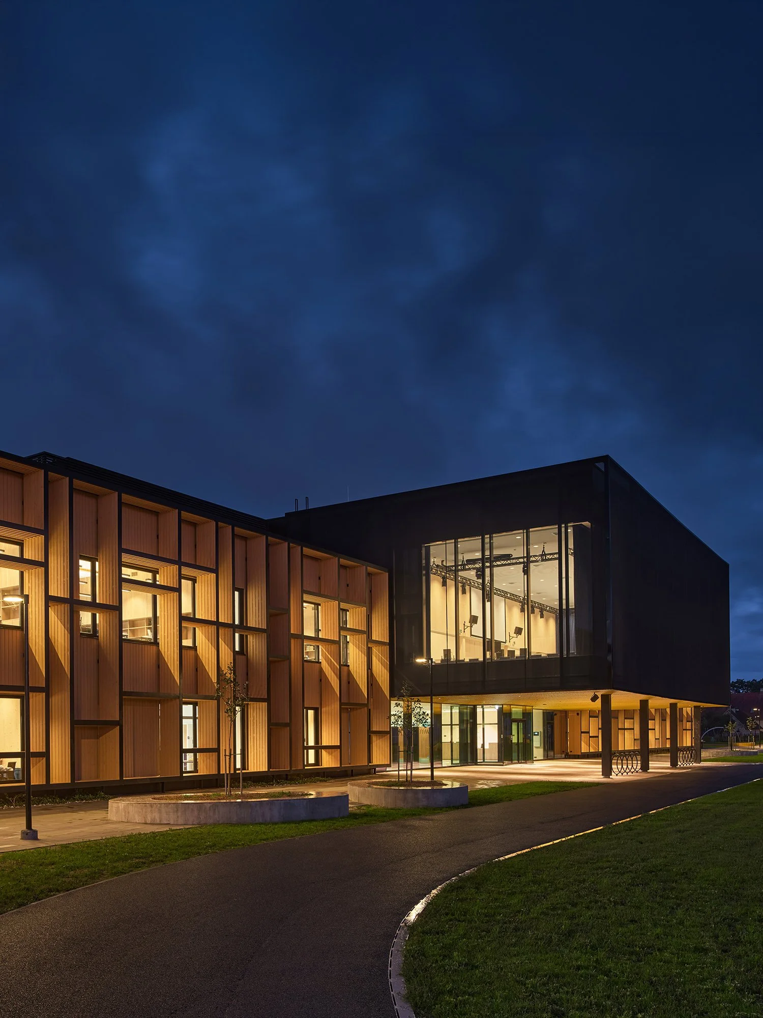 Modern building with wooden and black exterior, large glass windows on upper floor, warm interior lighting, surrounded by a sidewalk and small trees, against a dark evening sky.