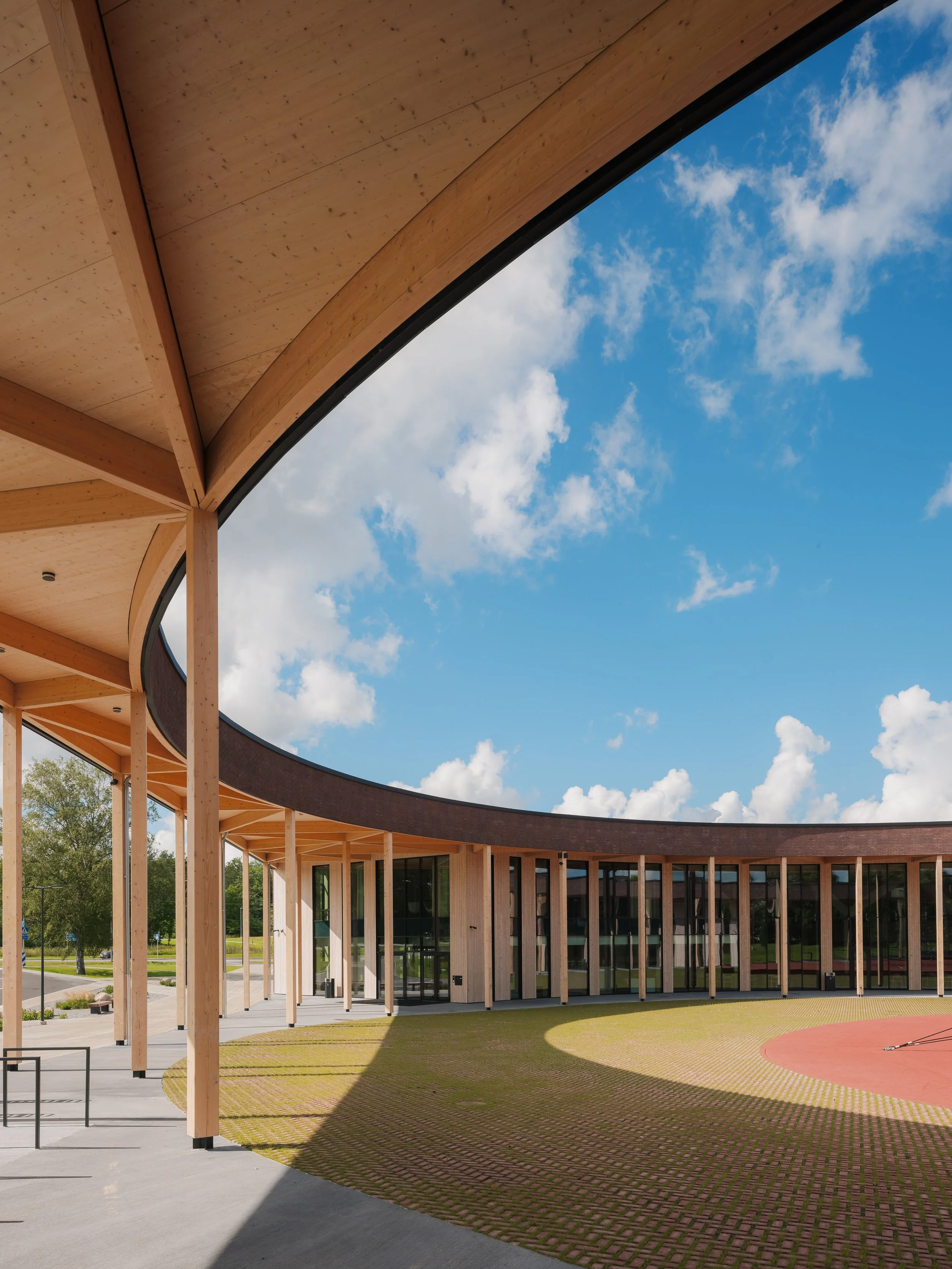Modern building with wooden supports and large glass windows, circular open courtyard with grass and a paved track, blue sky with white clouds.