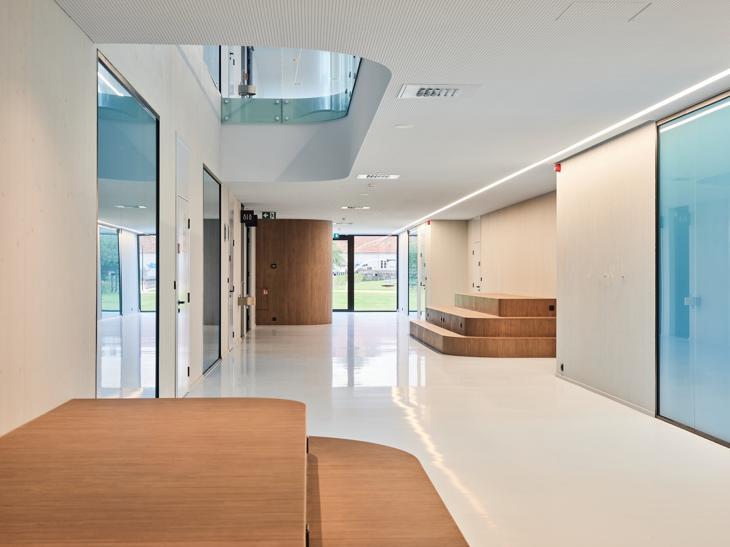 Modern, minimalist indoor space with white walls, wooden accents, and large glass doors and windows, providing a bright atmosphere.