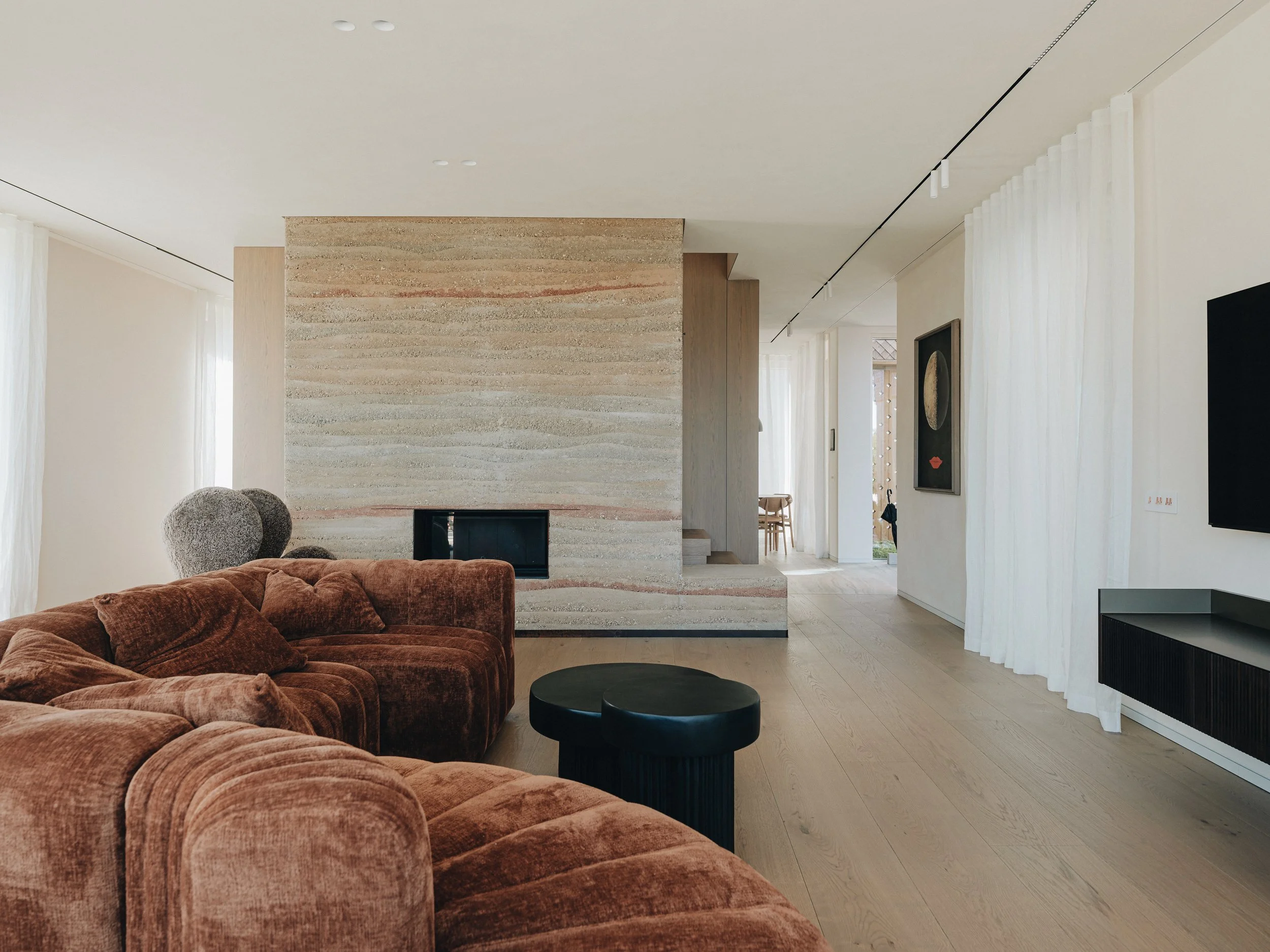 Modern living room with a large brown velvet sectional sofa, black round coffee table, and a beige stone fireplace. White curtains cover the windows, and artwork hangs on the walls. Light wood flooring and minimal decor complete the space.