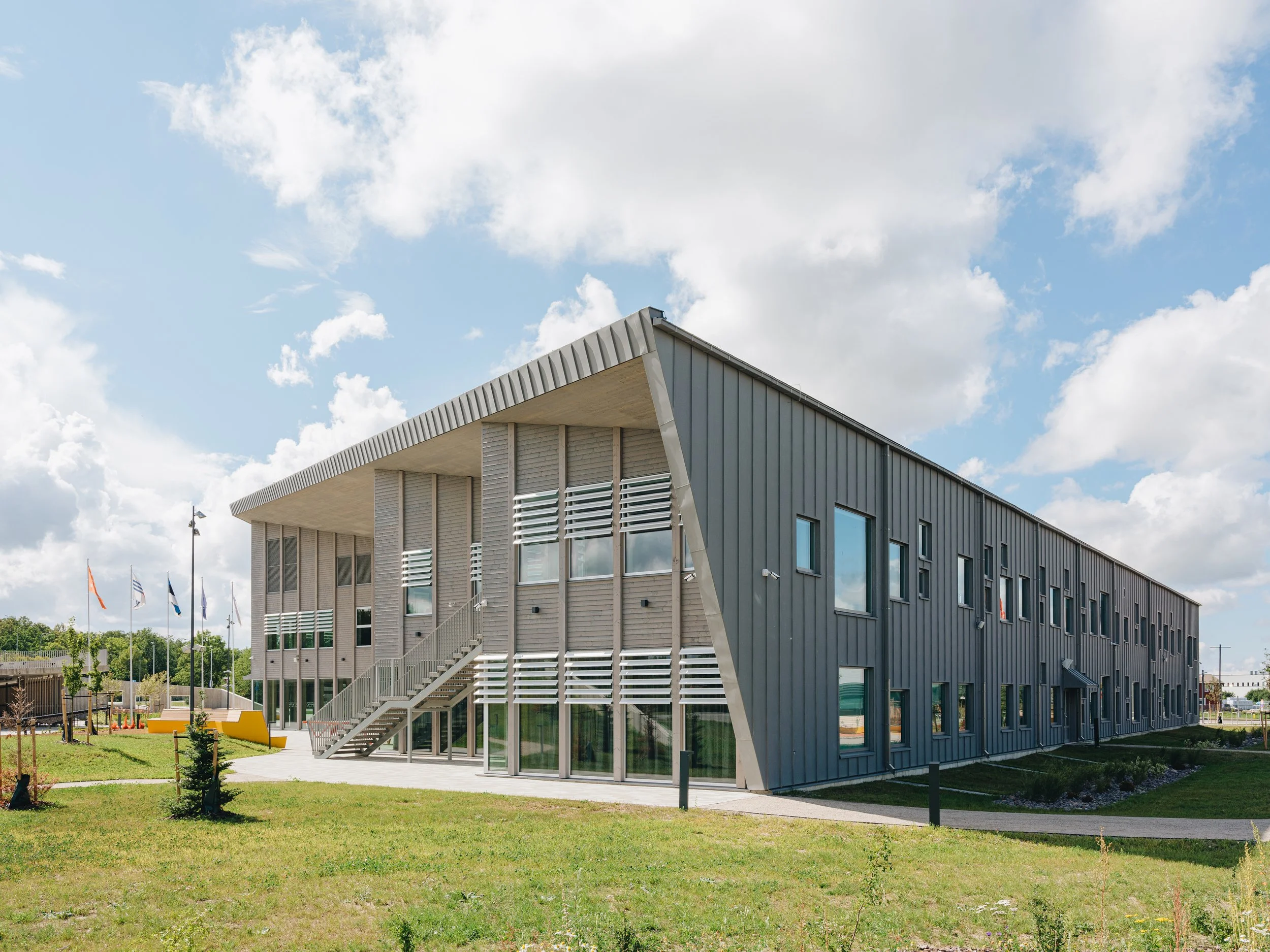 Modern two-story office building with gray metal siding, large windows, and an angled roof, situated in a grassy area with small trees and a pathway, under a partly cloudy sky.