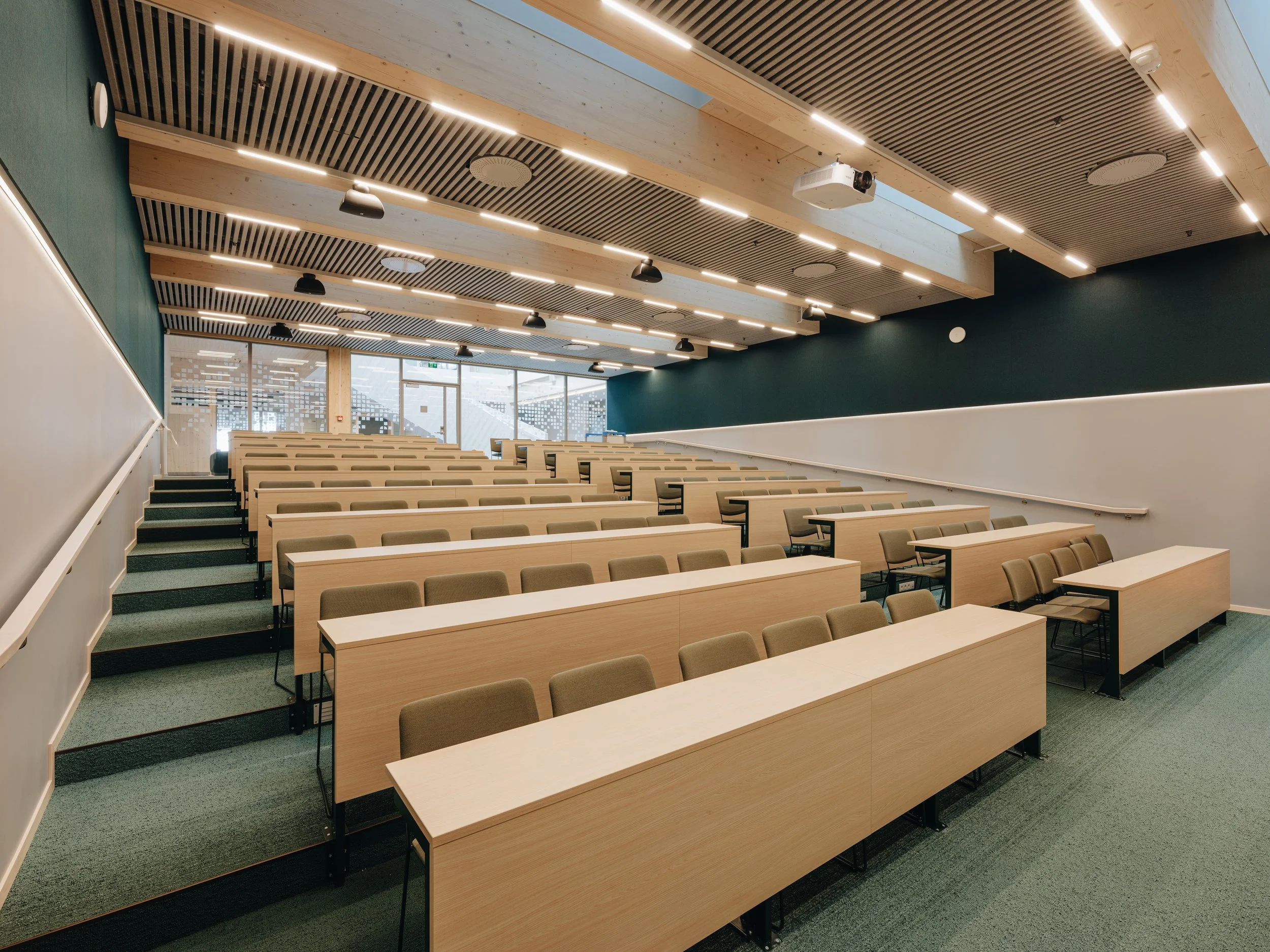 Empty university classroom with tiered seating, wooden desks, and chairs, large windows, and modern lighting.
