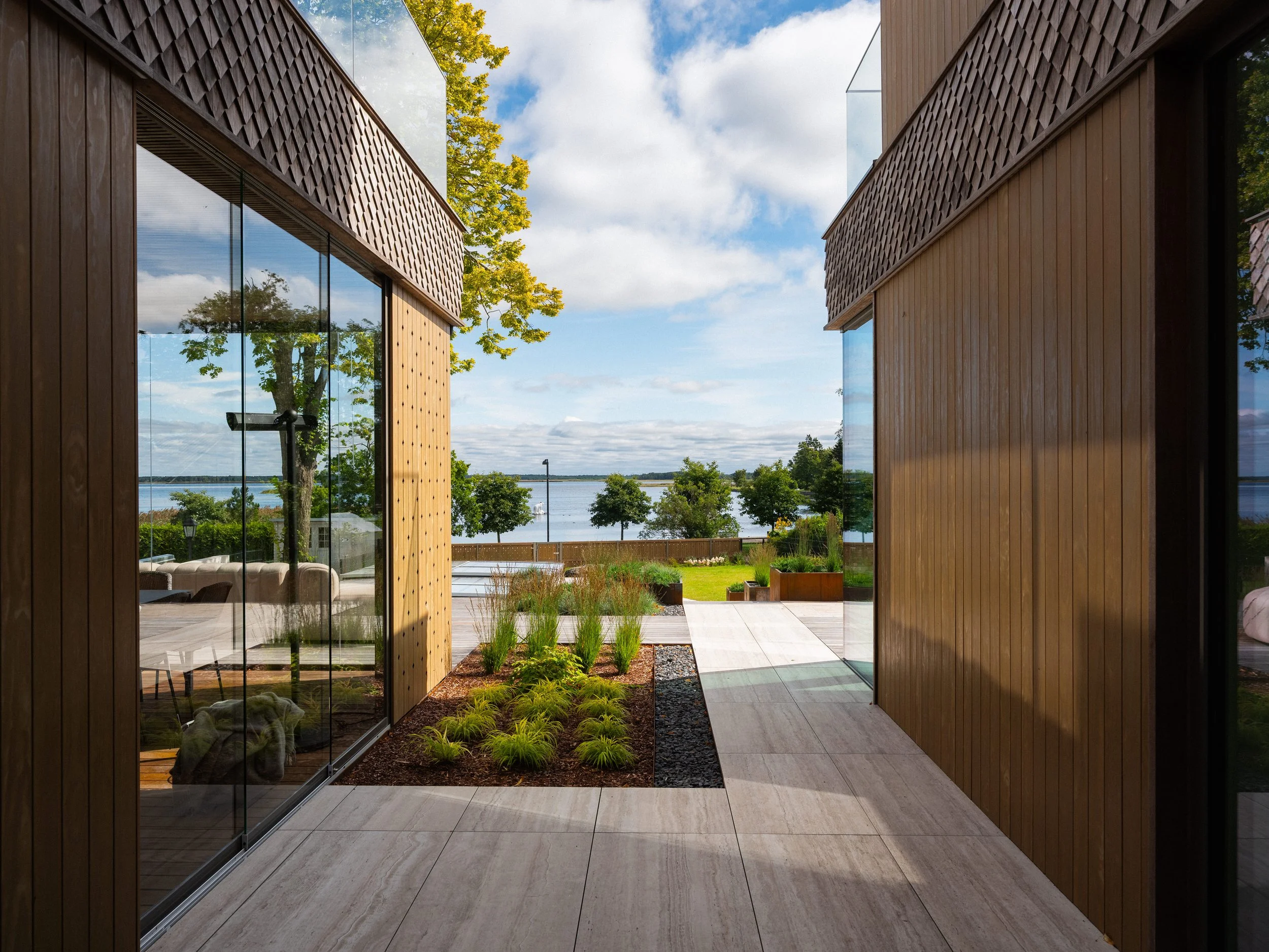 View through gap between two modern buildings with wooden and glass exteriors, overlooking a garden, a lake, trees, and a partly cloudy sky.