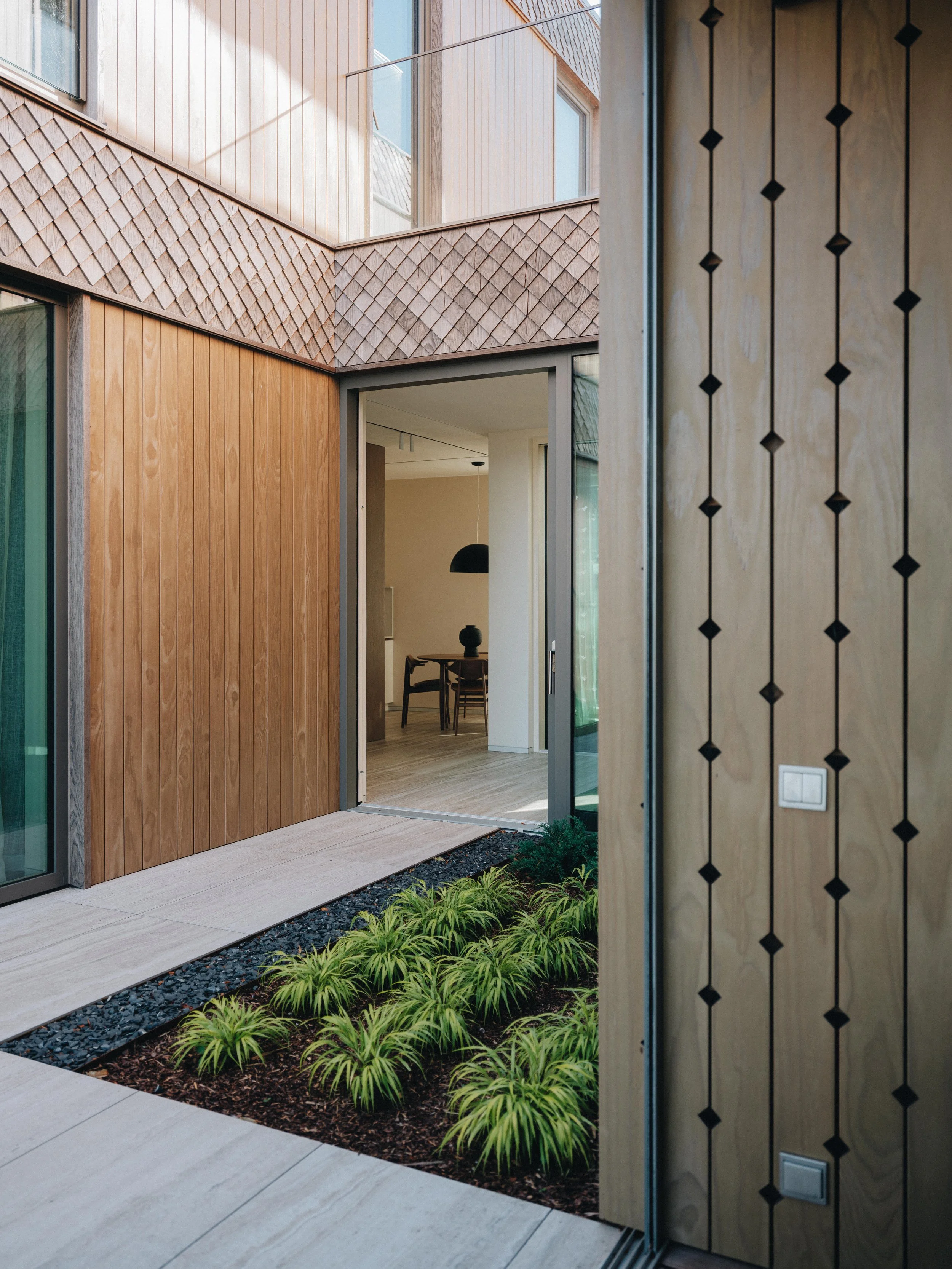 Modern home interior with a sliding glass door opening to a small garden area with green plants, wooden and textured wall panels, and a dining room with a black pendant light visible inside.