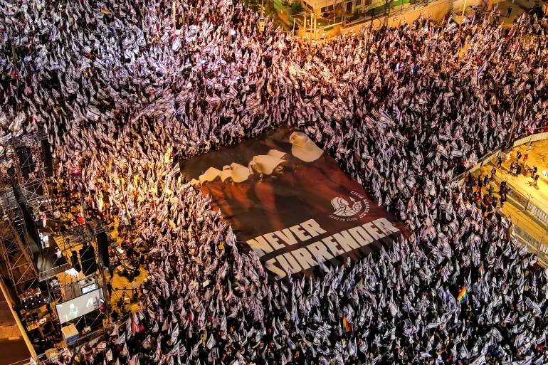 A large crowd of people holding up numerous white flags surrounds a giant banner that reads "NEVER SURRENDER" with a logo and patriotic imagery, in an outdoor stadium or arena at night.