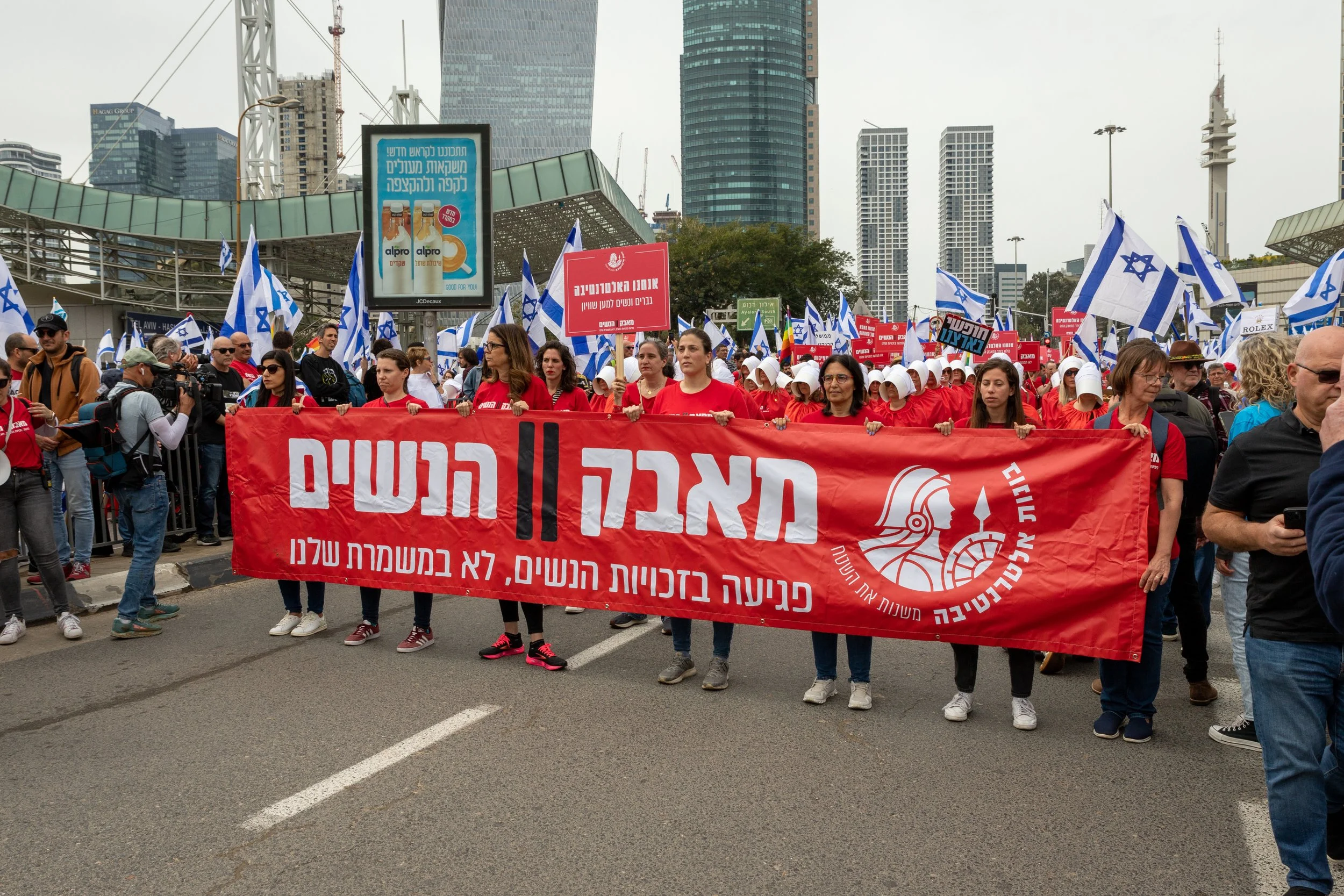 A large group of people participating in a protest march in an urban area, holding a red banner with Hebrew text. Many also carry Israeli flags and wear red shirts. The background features modern high-rise buildings and billboards.